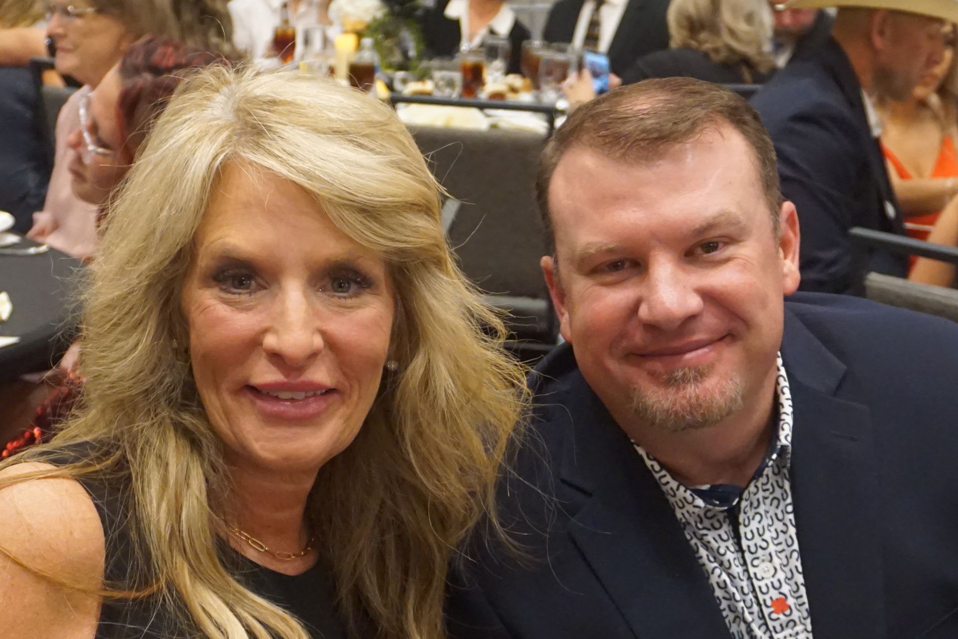 A smiling couple sits together at a table in a dimly lit, crowded event space.