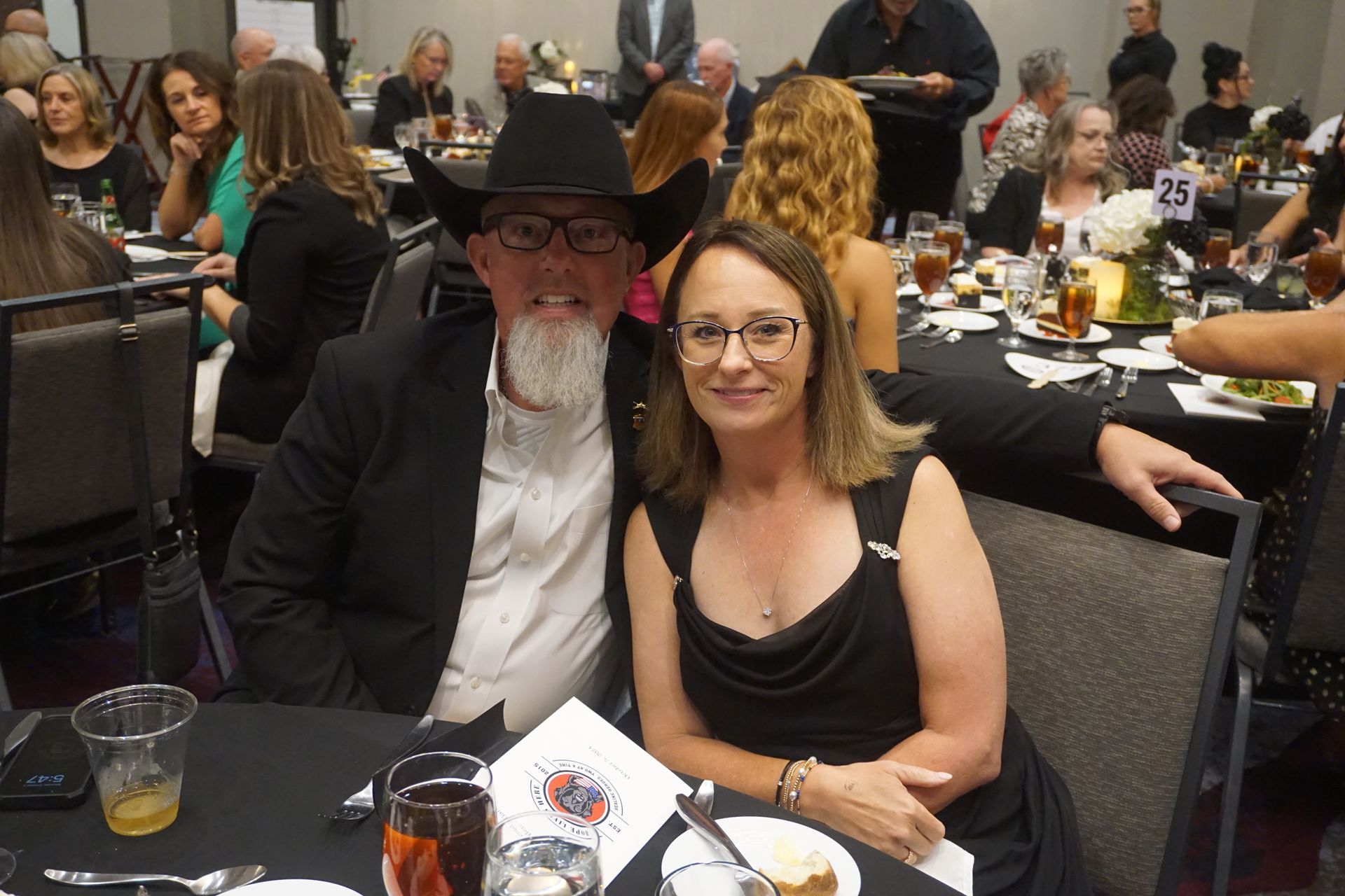 A couple sits at a table in a banquet hall, smiling at the camera during a formal event.