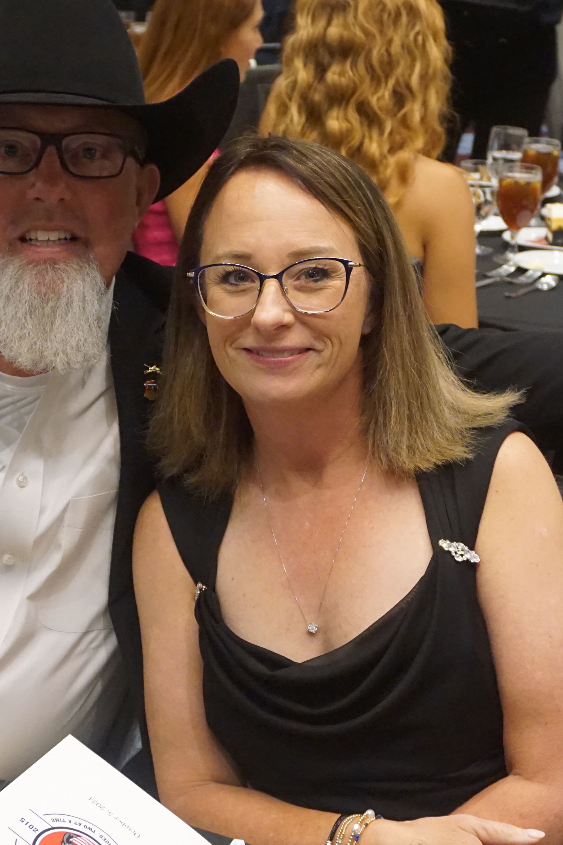 A person with light brown hair wearing glasses and a black dress sits next to a person in a cowboy hat at a banquet.