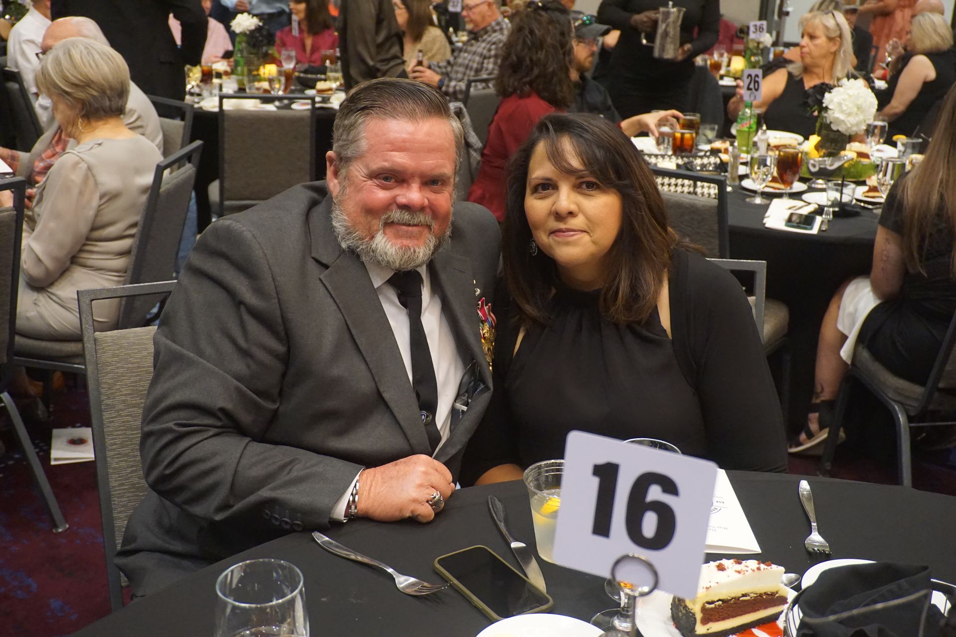 A man and woman pose at a table marked with the number 16 in a banquet hall setting.