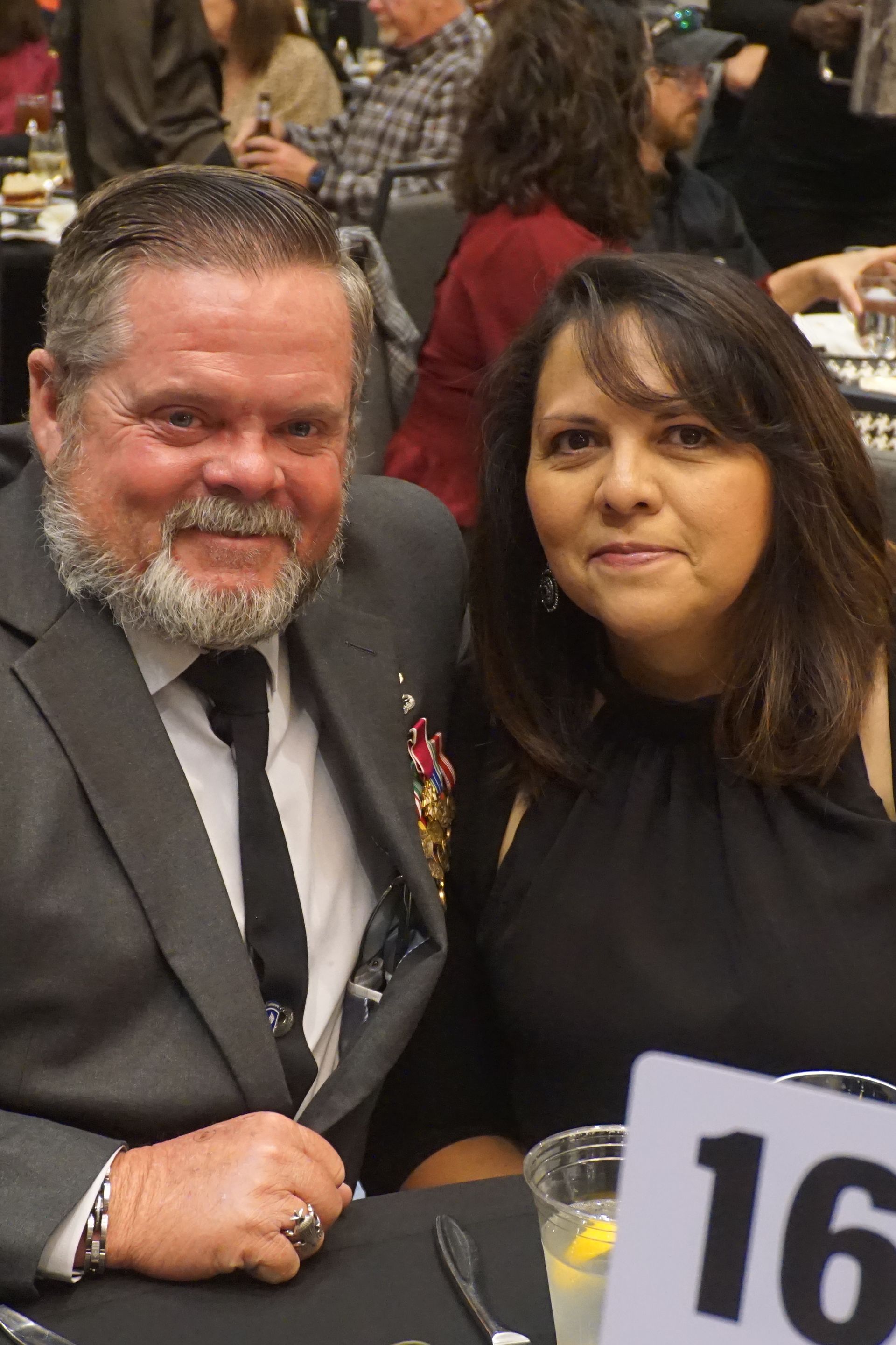 A man in a suit and tie and a woman in a black dress posing together at a table, with a number 16 sign visible.
