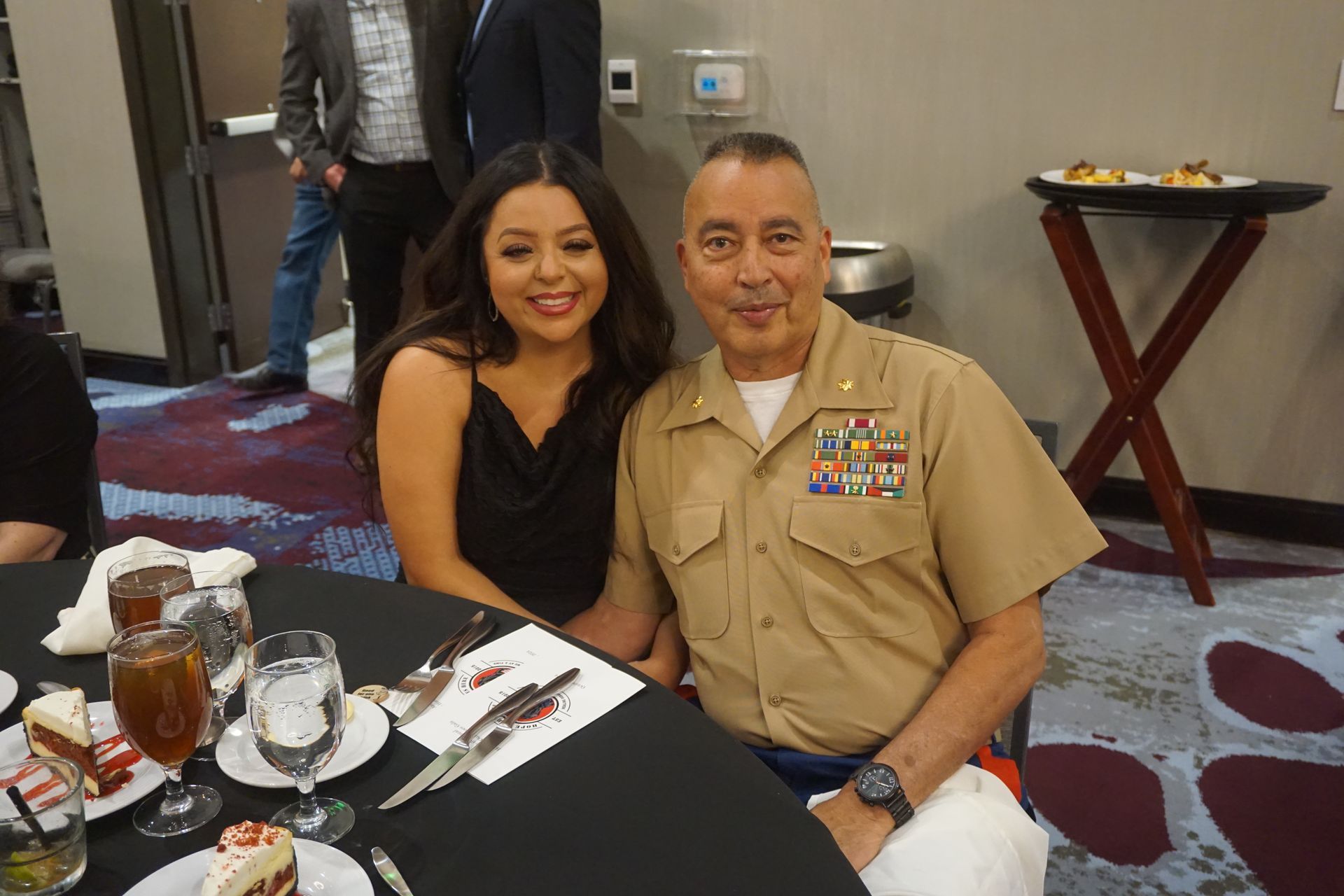 A smiling woman and a person in a tan military uniform sit together at a formal table with drinks and cake.