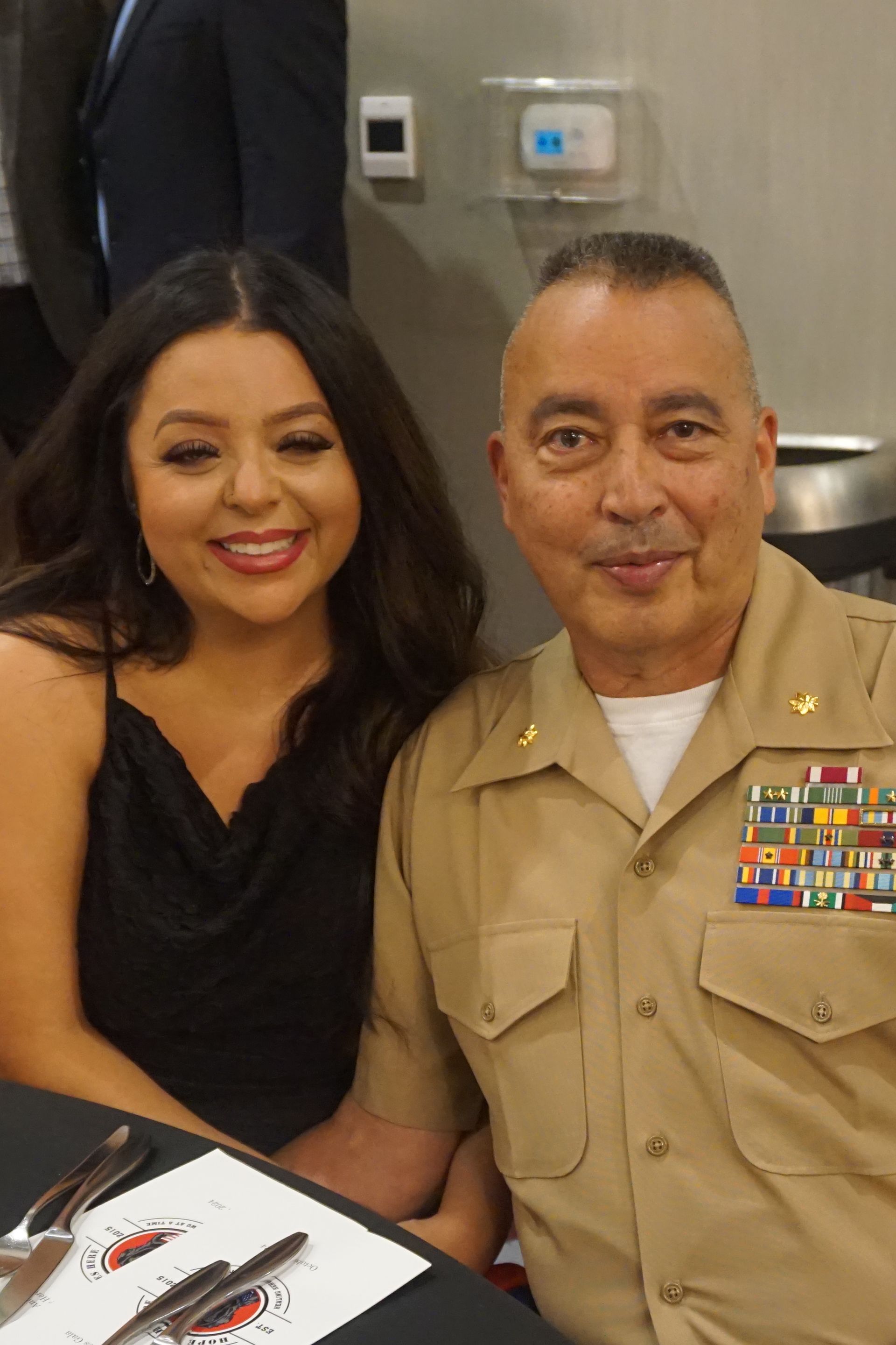A person in a tan military uniform with a ribbon rack sits next to a person in a black top at a table with silverware.