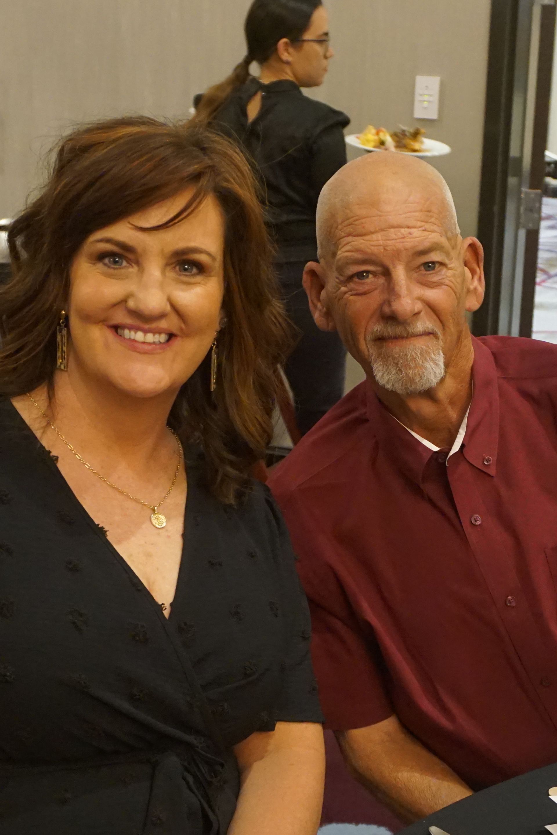 A smiling woman and a man with a goatee in a burgundy shirt sitting together in an indoor event space.