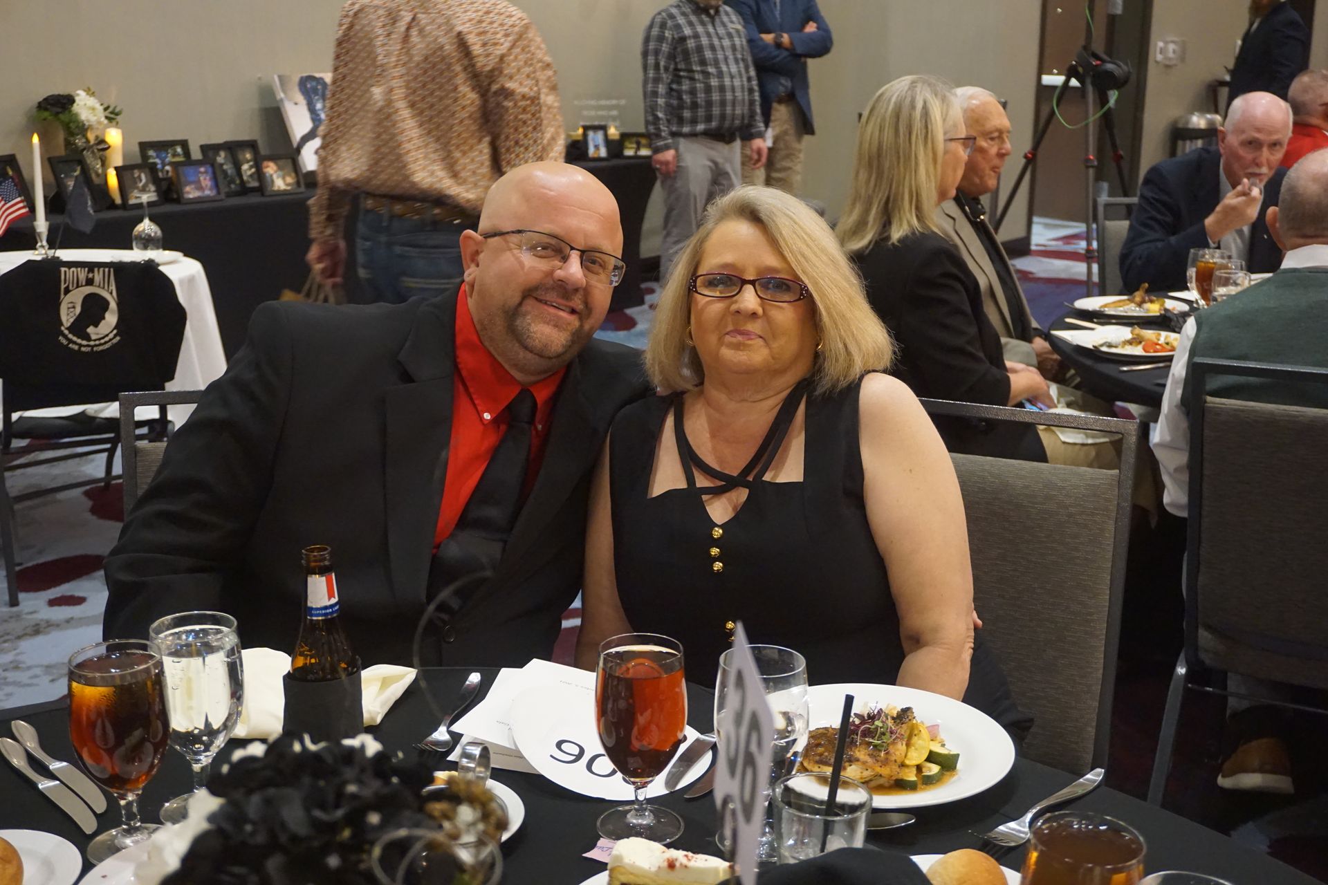 A couple smiles at a table during a banquet, featuring place settings and guests in the background.