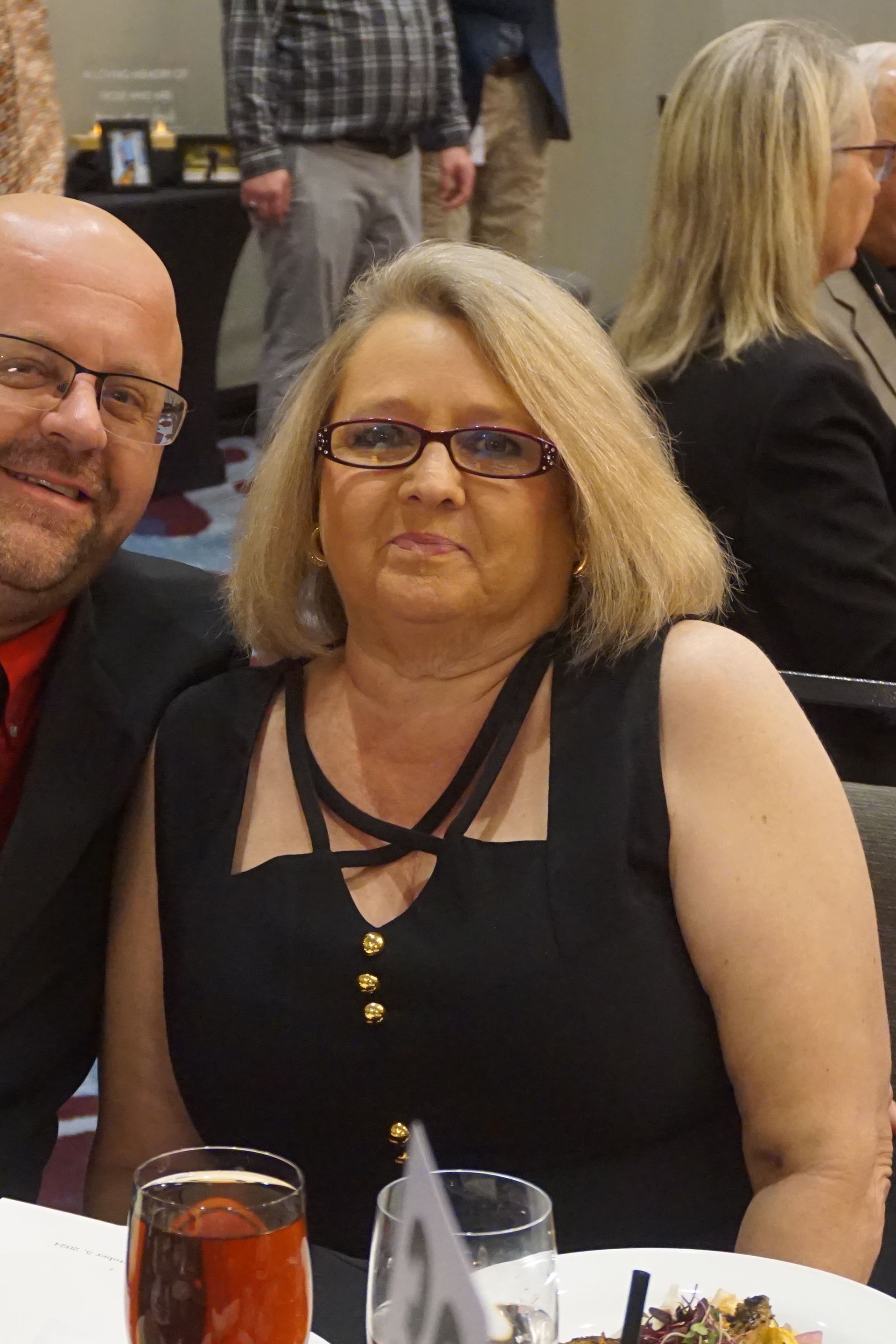 A smiling man and a woman in a black sleeveless dress sit together at a table in a dimly lit event space.