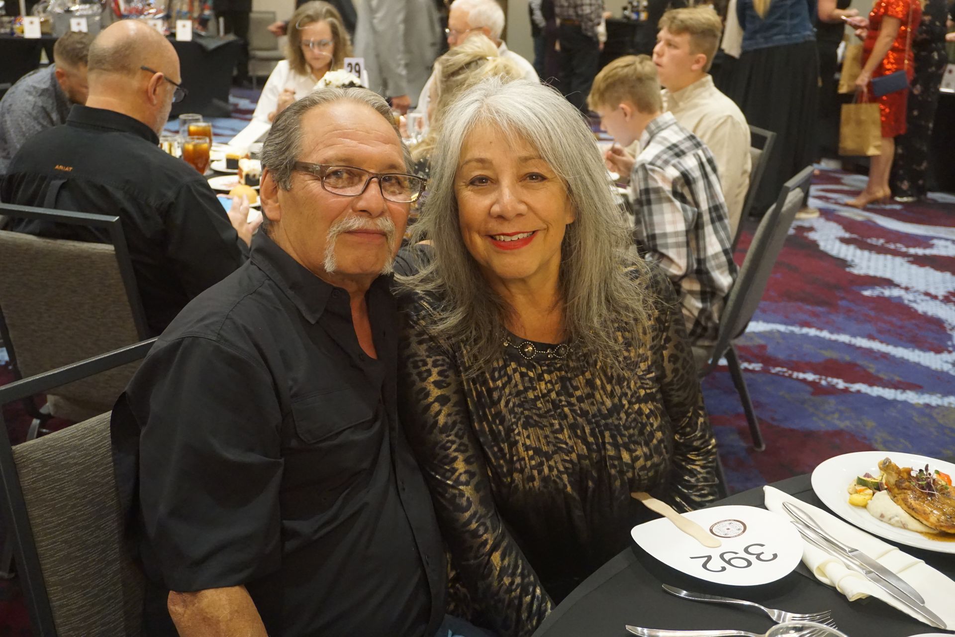 A smiling couple sits at a table in a banquet hall, with other guests visible in the blurred background.