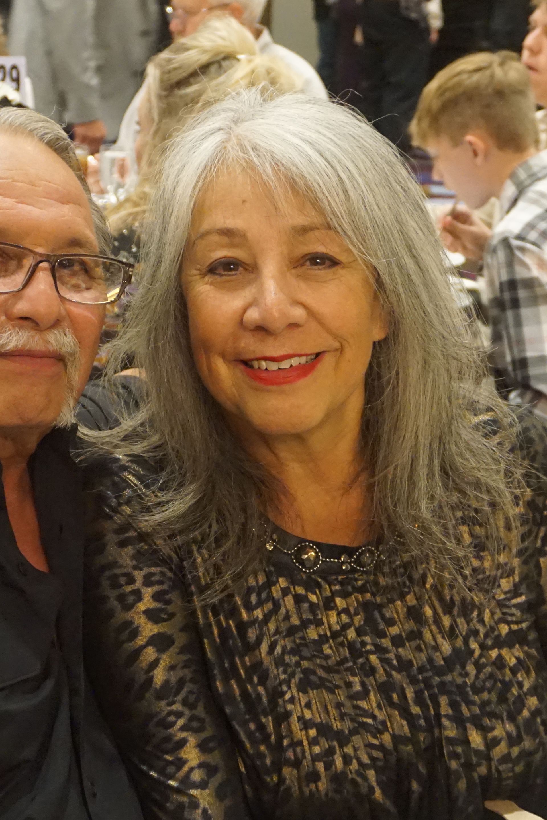 A smiling person with long gray hair in a black animal-print top poses next to another individual at an indoor event.