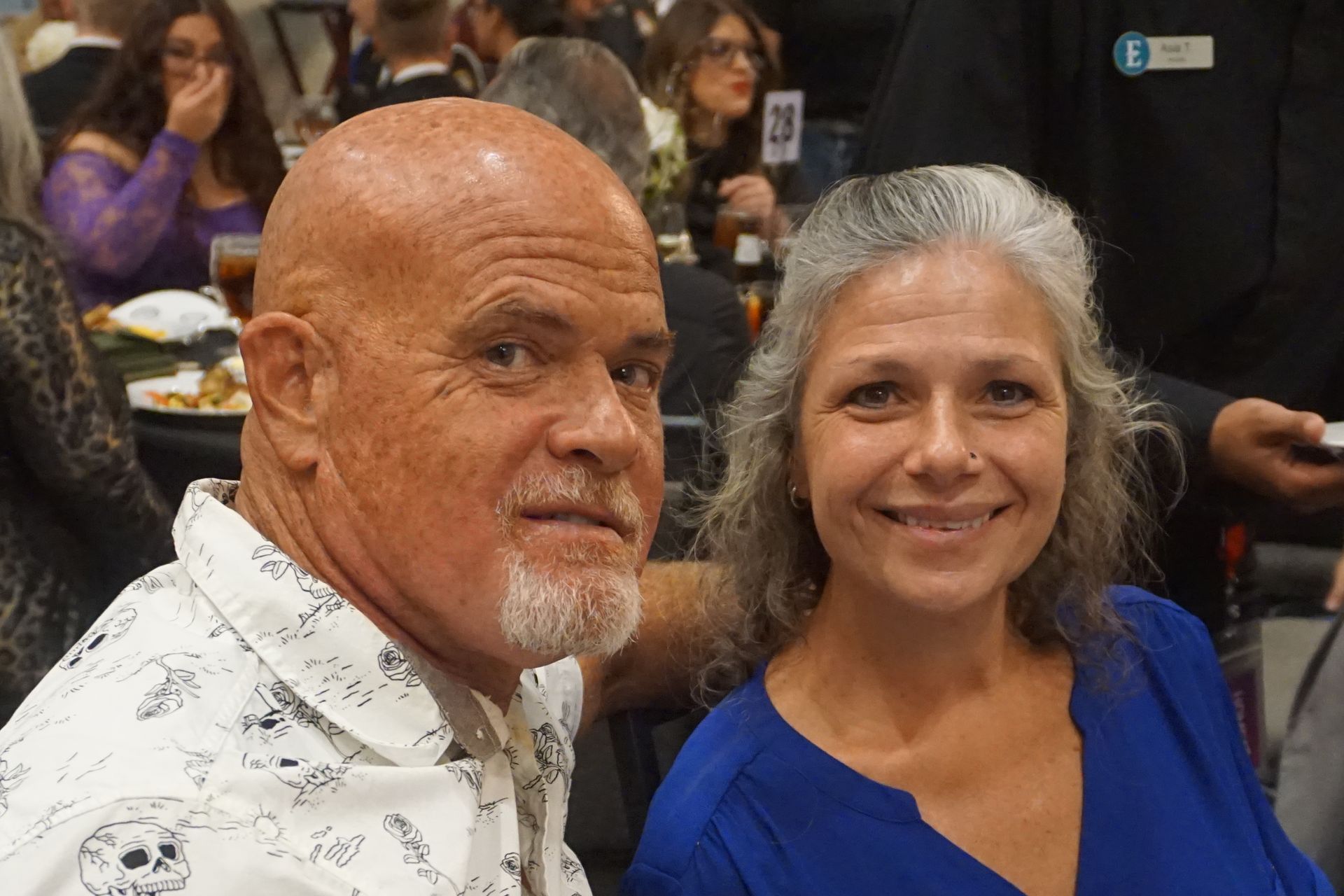 A bald man in a white skull-print shirt and a woman in a blue blouse smile closely together at a social event.