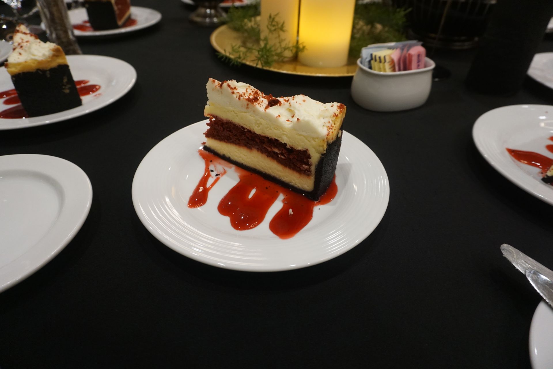 A slice of red velvet cheesecake drizzled with red fruit sauce on a white plate, set on a dark tablecloth.