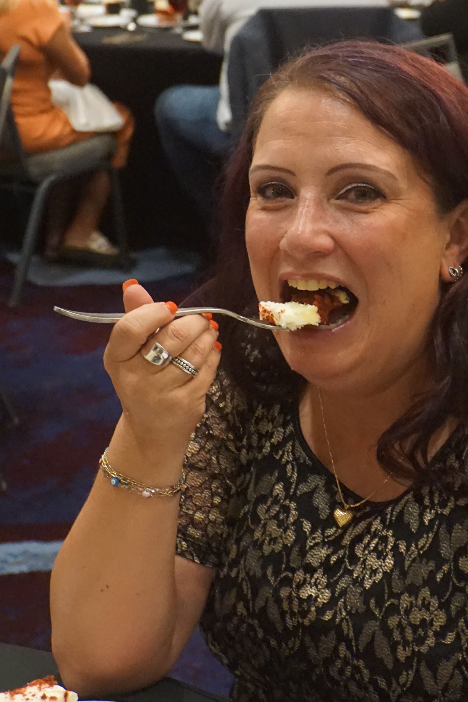 A person wearing a black lace top eats a piece of dessert with a silver fork at a table in a room.