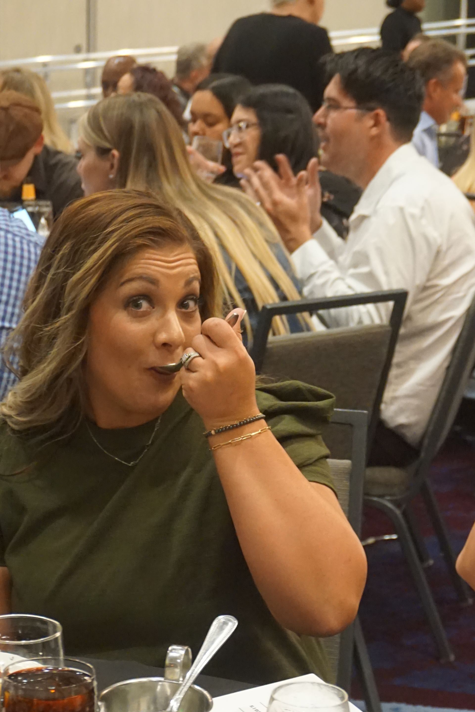 A woman in an olive green top smiles with a spoon to her mouth in a crowded event hall, others dining in the background.