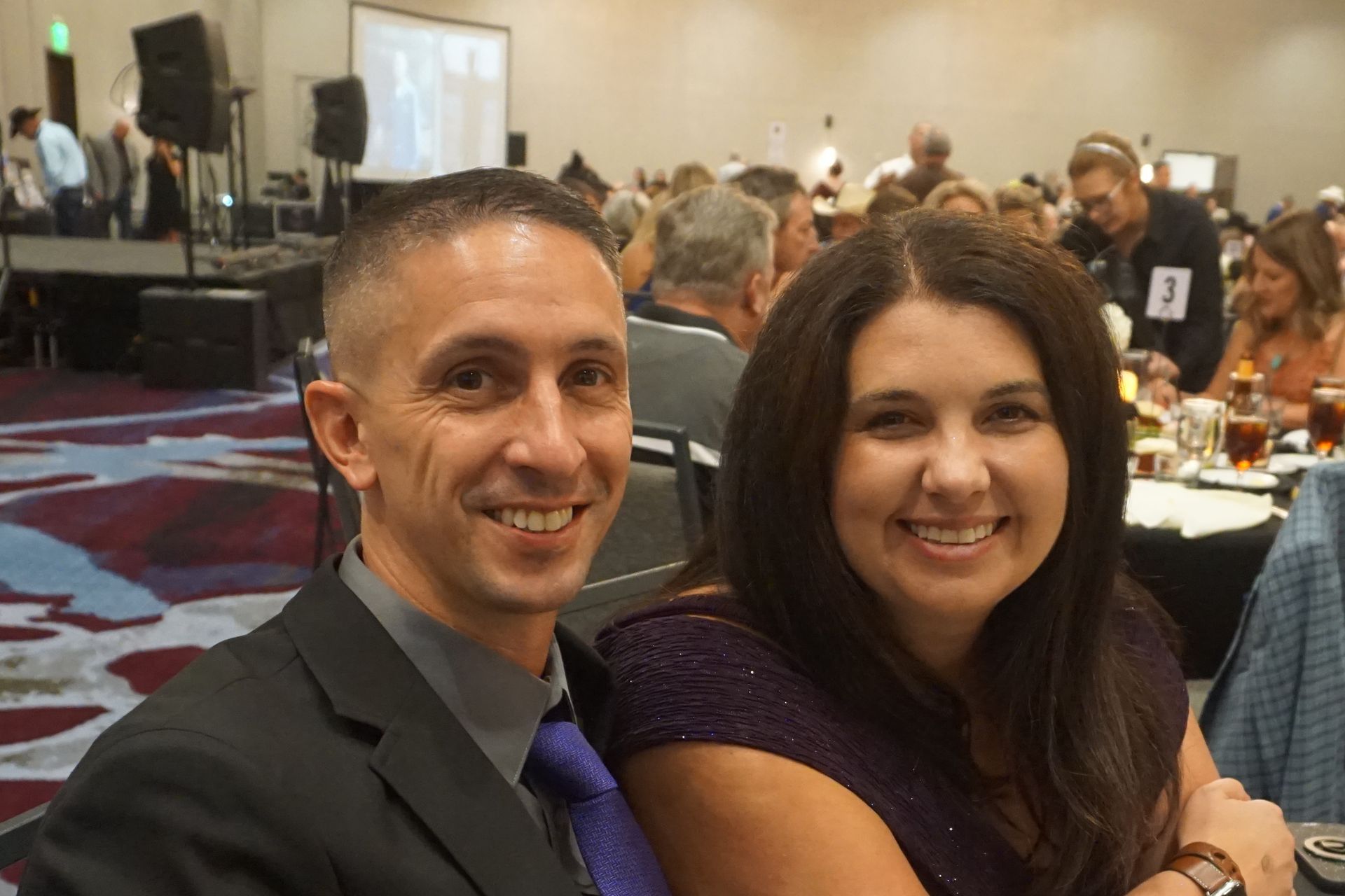 A smiling couple in formal attire poses together at a brightly lit event in a large ballroom.
