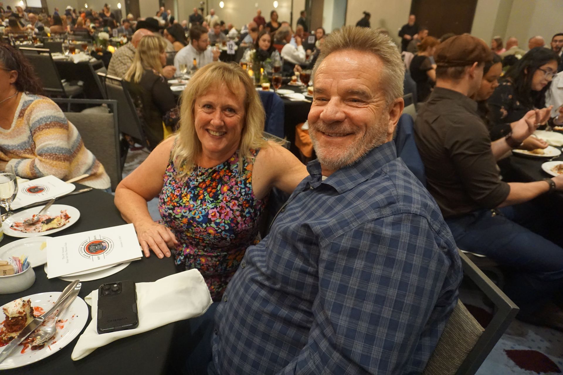 A couple smiles at a banquet table in a large, crowded event space.