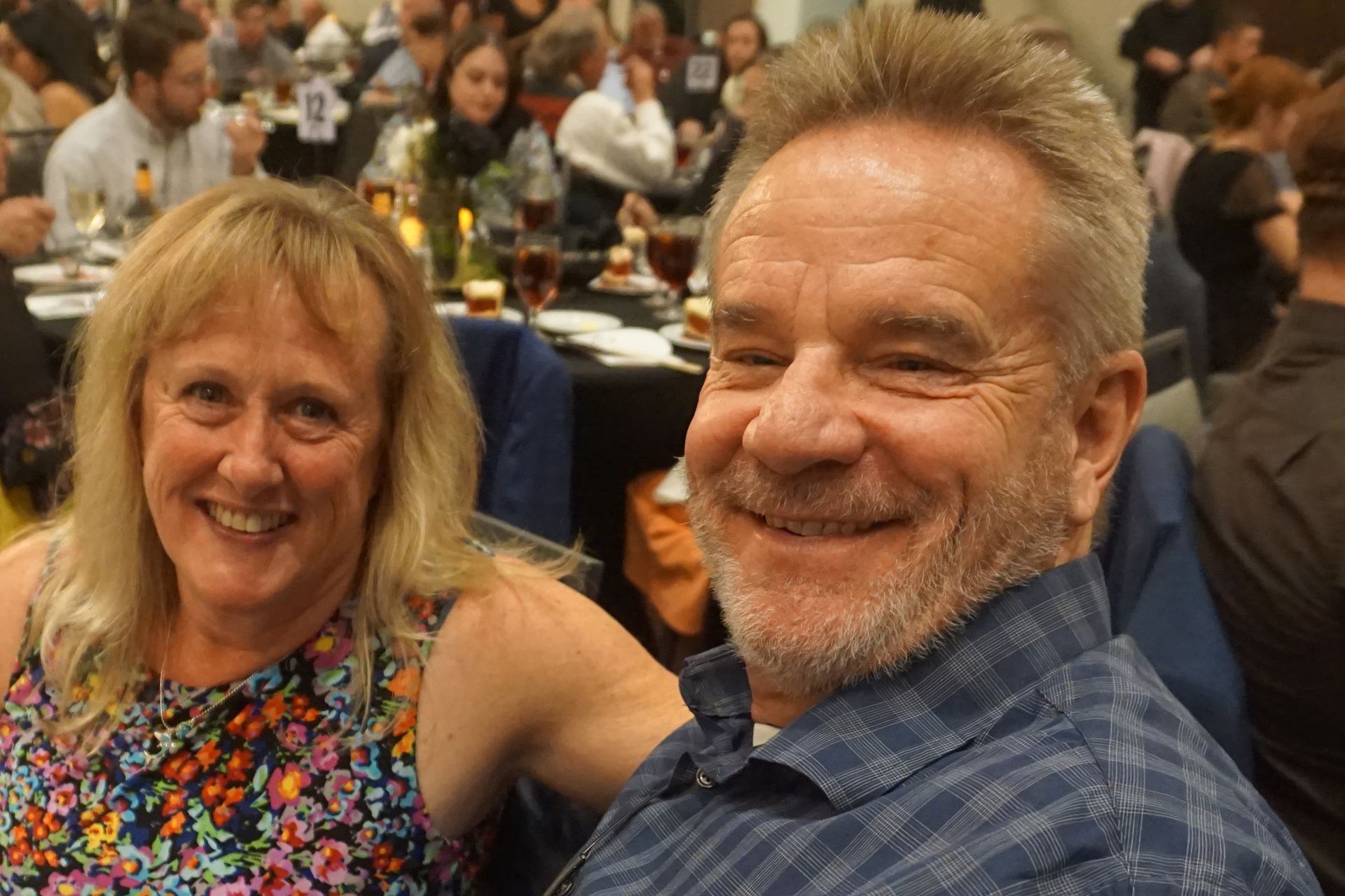 A smiling couple posing for a selfie at a banquet, with guests and tables visible in the blurred background.
