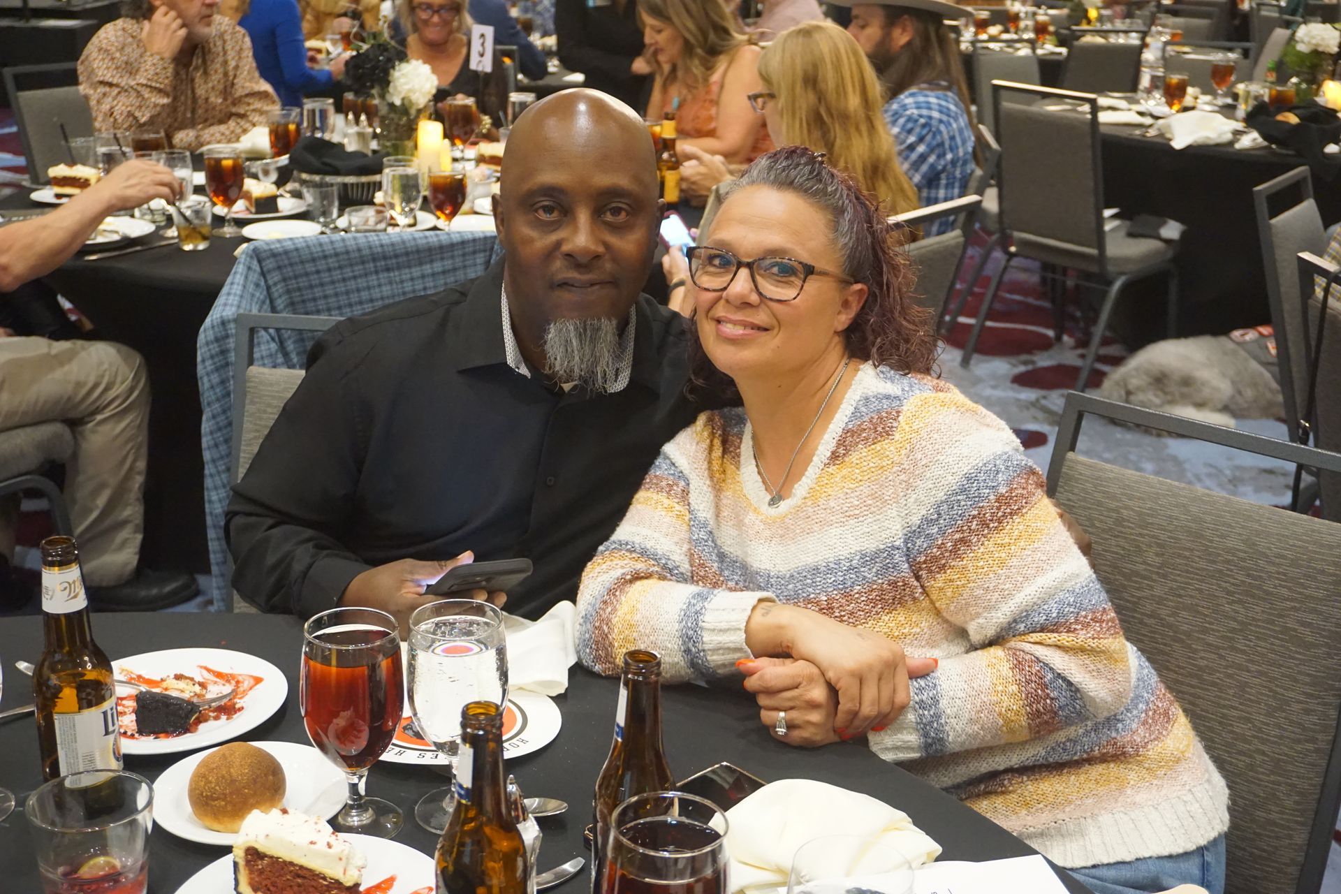 A couple sits at a table in a dimly lit banquet hall, smiling for the camera during a social event.