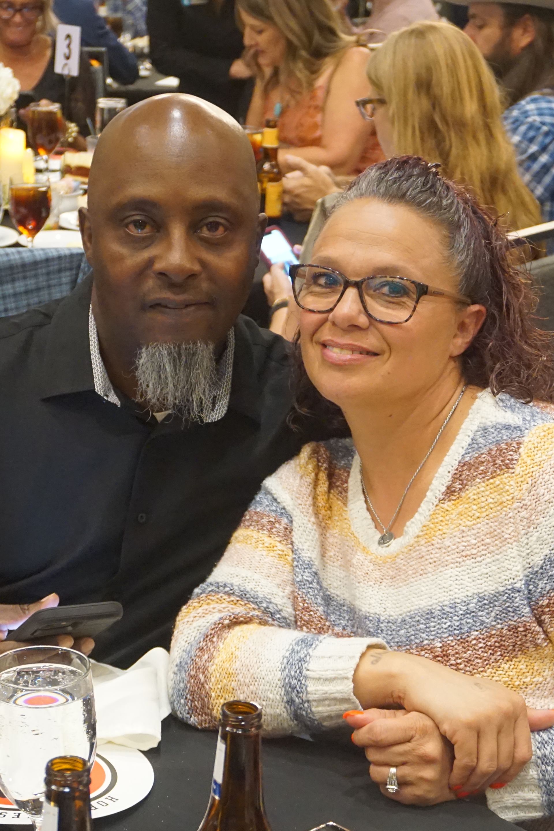 A couple poses for a photo at a table in a restaurant, both smiling at the camera.