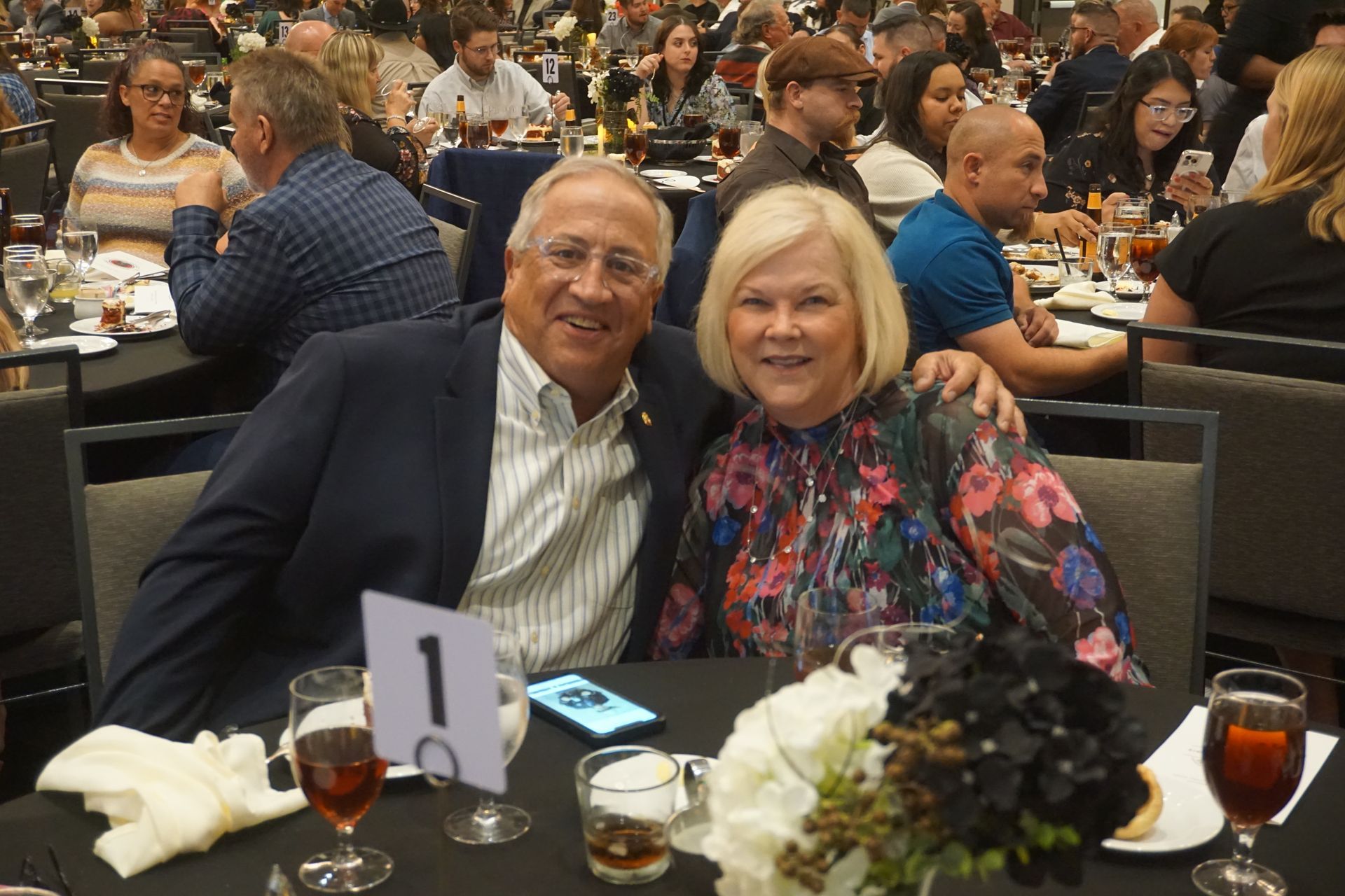 An older couple smiling together at a table marked with the number one at a banquet, with guests seated in the background.