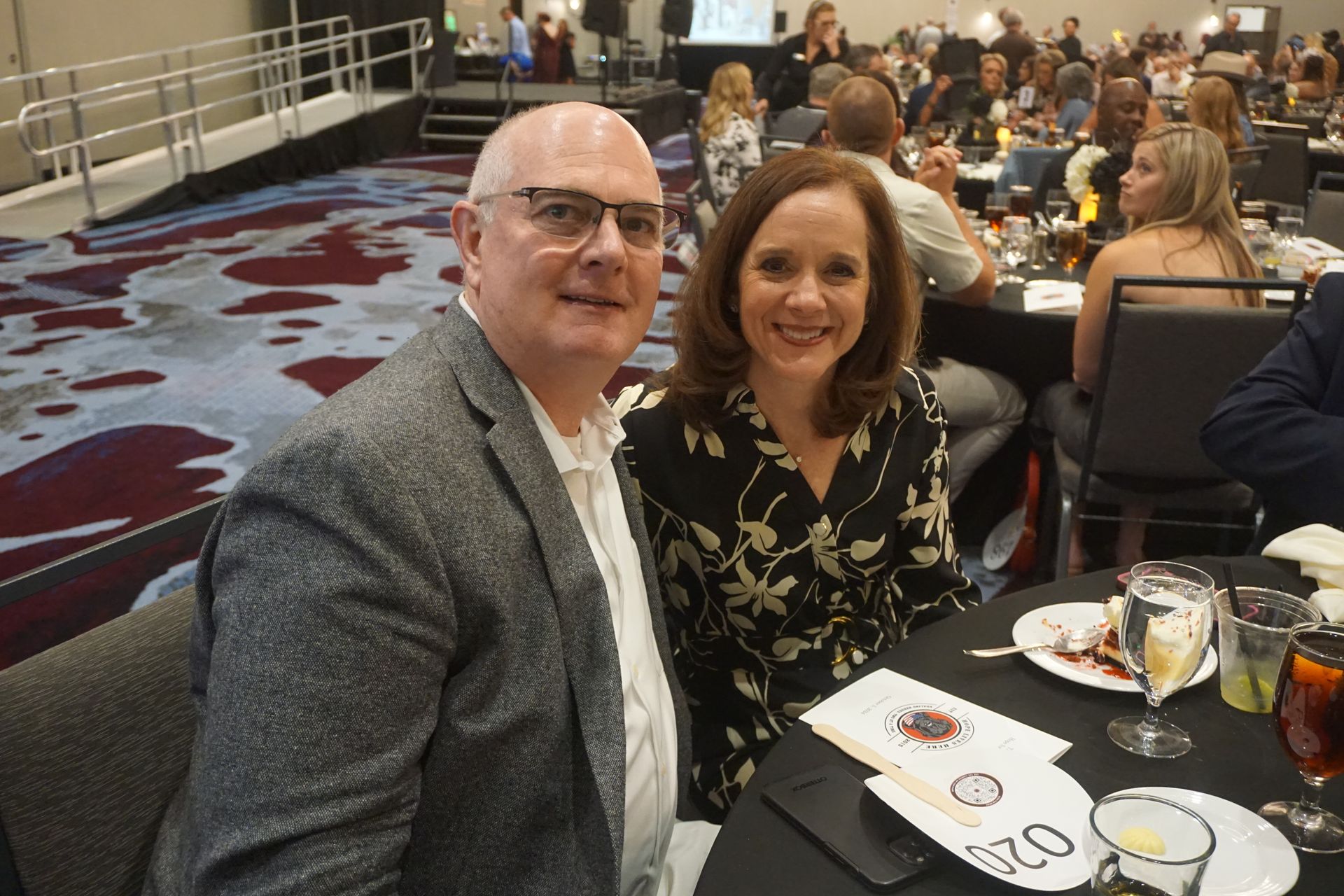 A man and woman sit at a round table at an event, smiling at the camera in a room filled with seated guests.