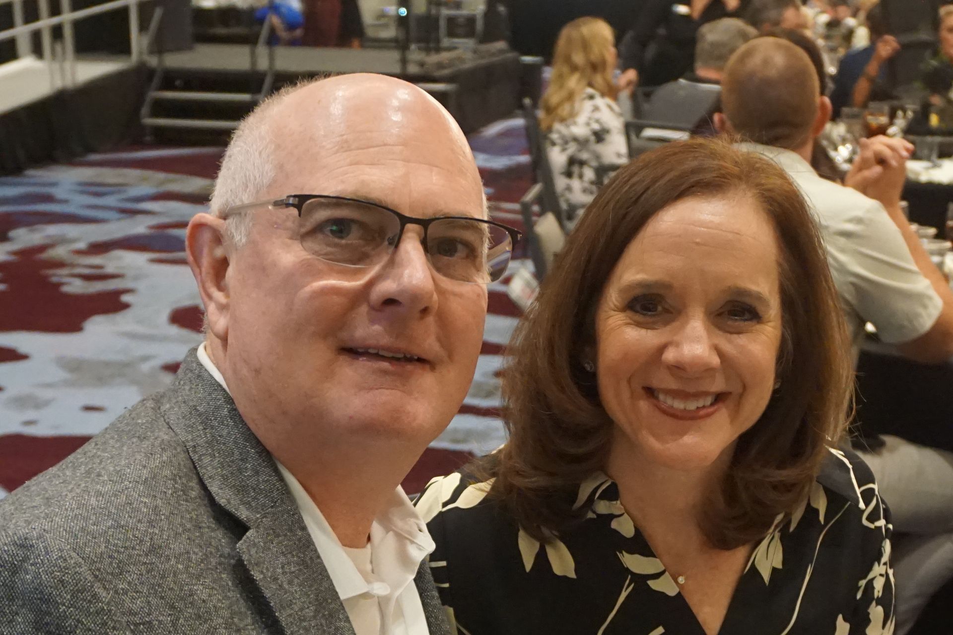 A smiling man in glasses and a blazer stands next to a smiling woman in a black and white floral top at an indoor event.