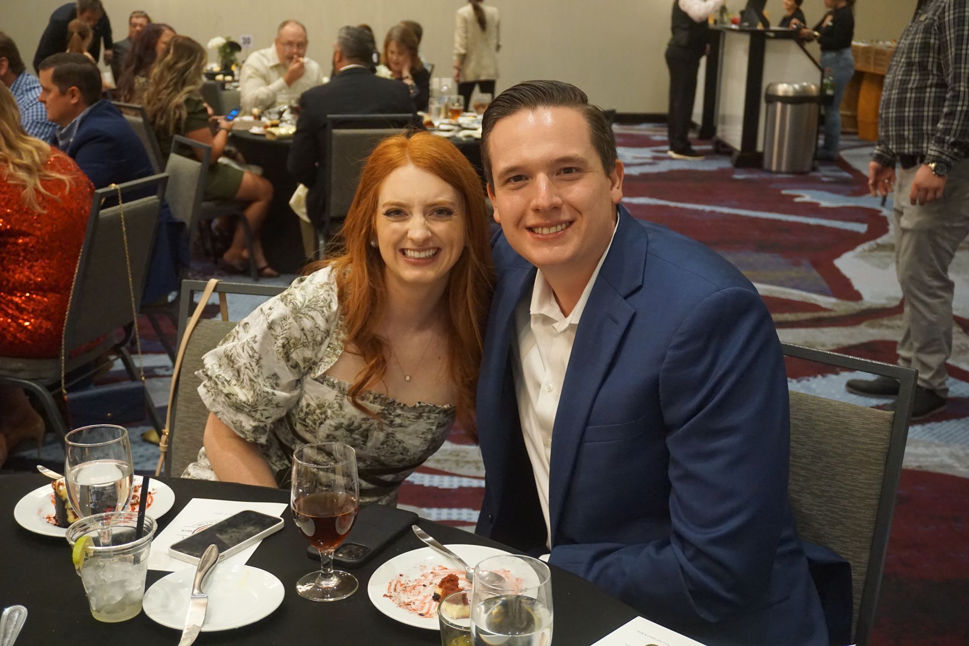 A smiling couple sits at a table during an event, posing for a photo in a room with other guests in the background.