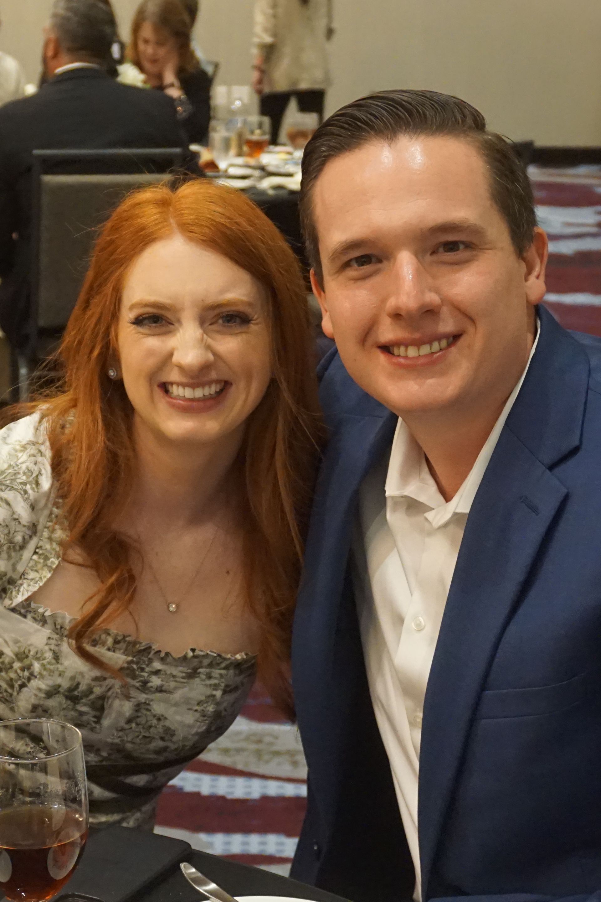 A smiling man in a blue blazer and a woman with red hair in a patterned dress pose together at a table in a banquet hall.