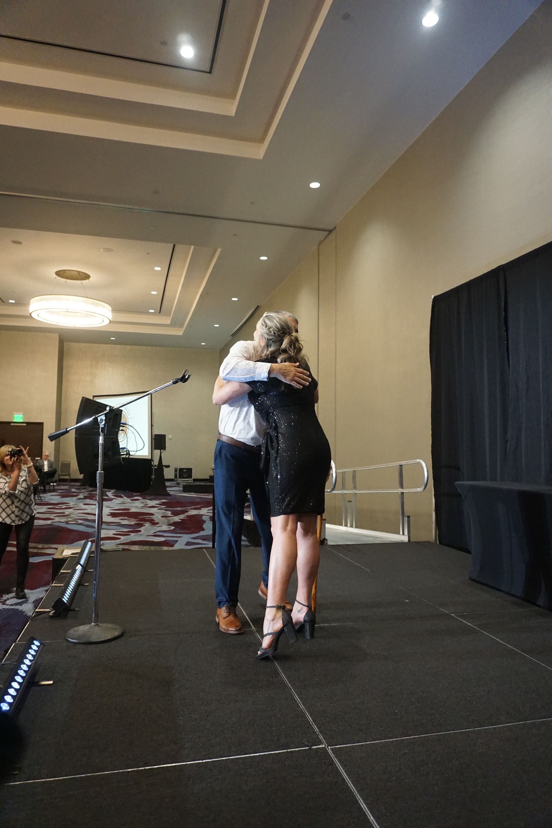 A person in a black dress and a person in a white shirt hug on a stage in a hotel ballroom.