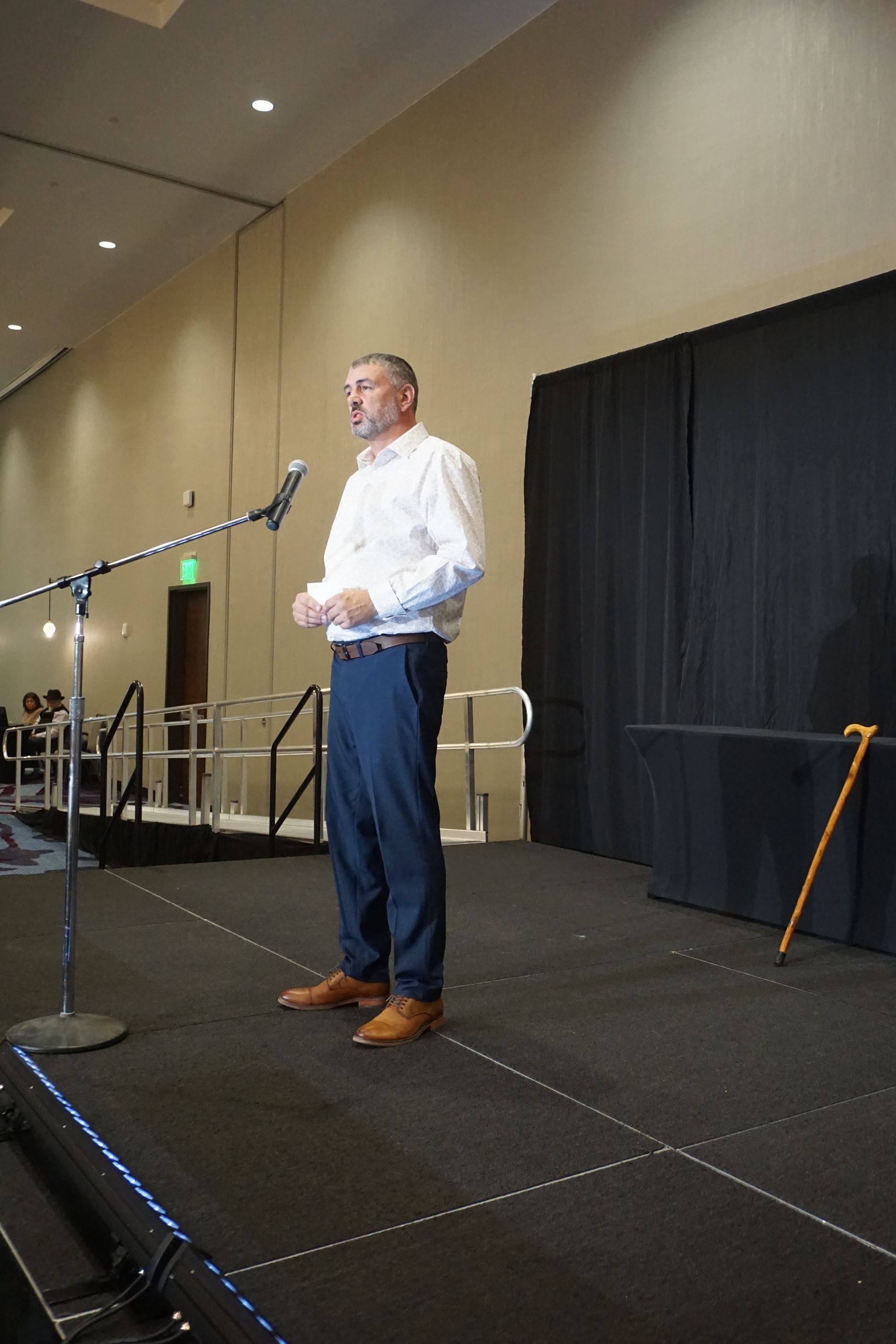 A speaker stands at a microphone on a stage with a cane resting against a table nearby.