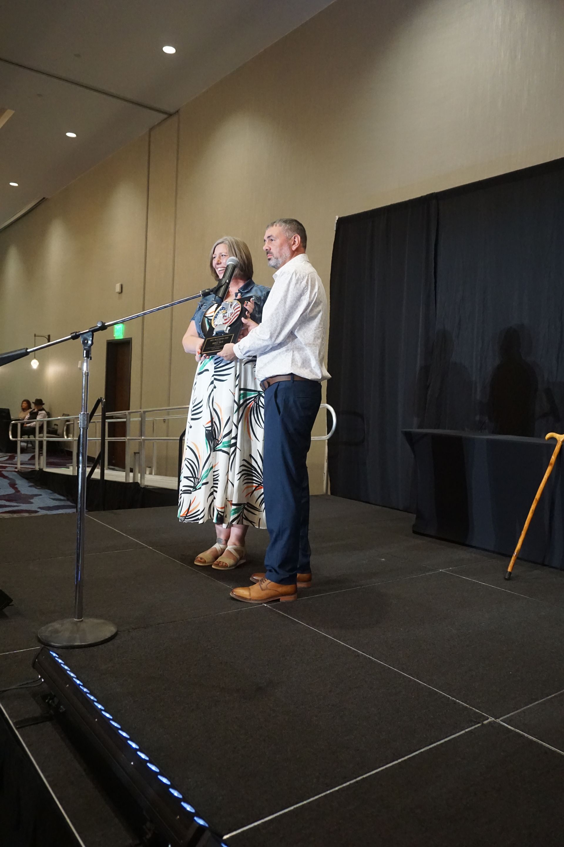 A person in a floral skirt and denim jacket stands on stage with a person in a white button-down shirt at a microphone.
