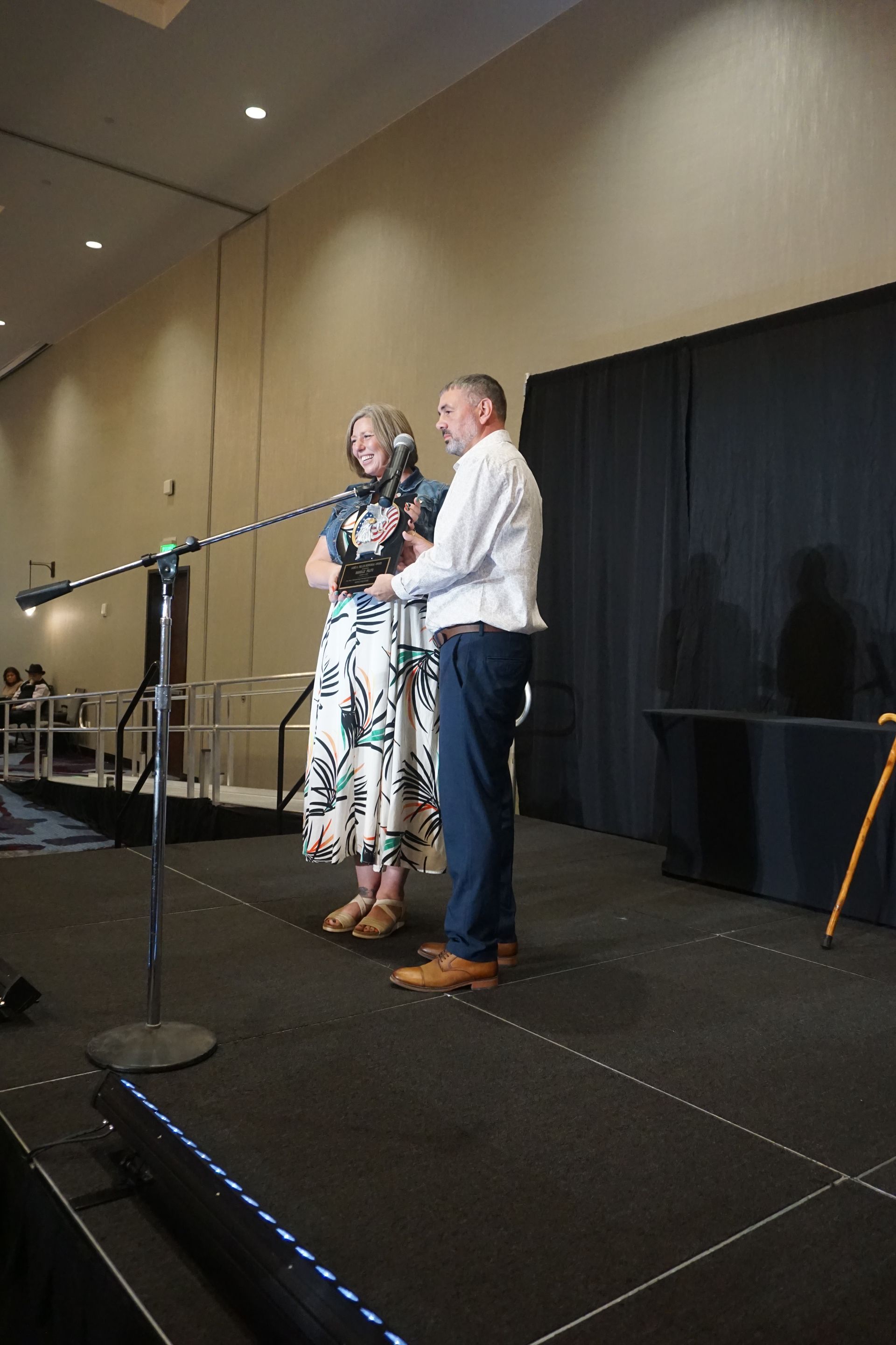 Two people stand on a stage with a microphone, presenting an object to an audience in a conference room.