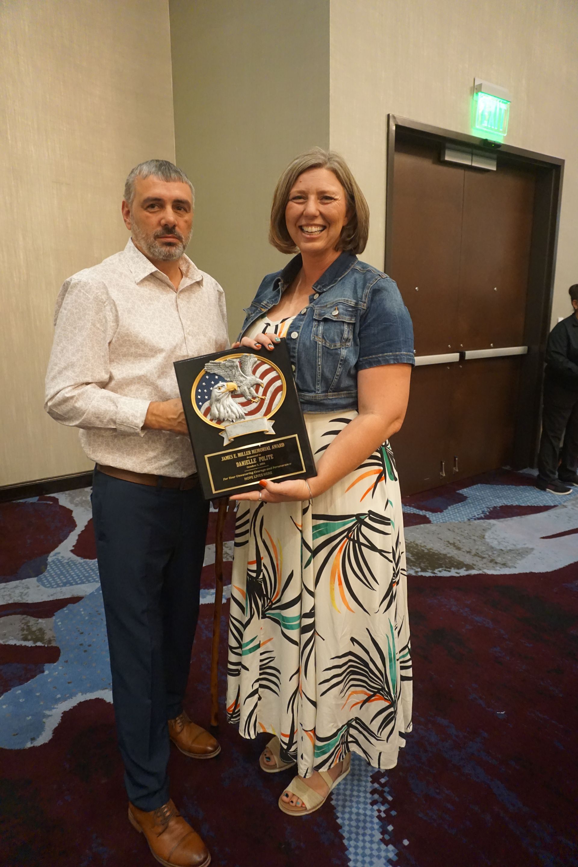 A person in a light shirt and dark trousers presents an award to a person in a jean jacket and patterned dress.