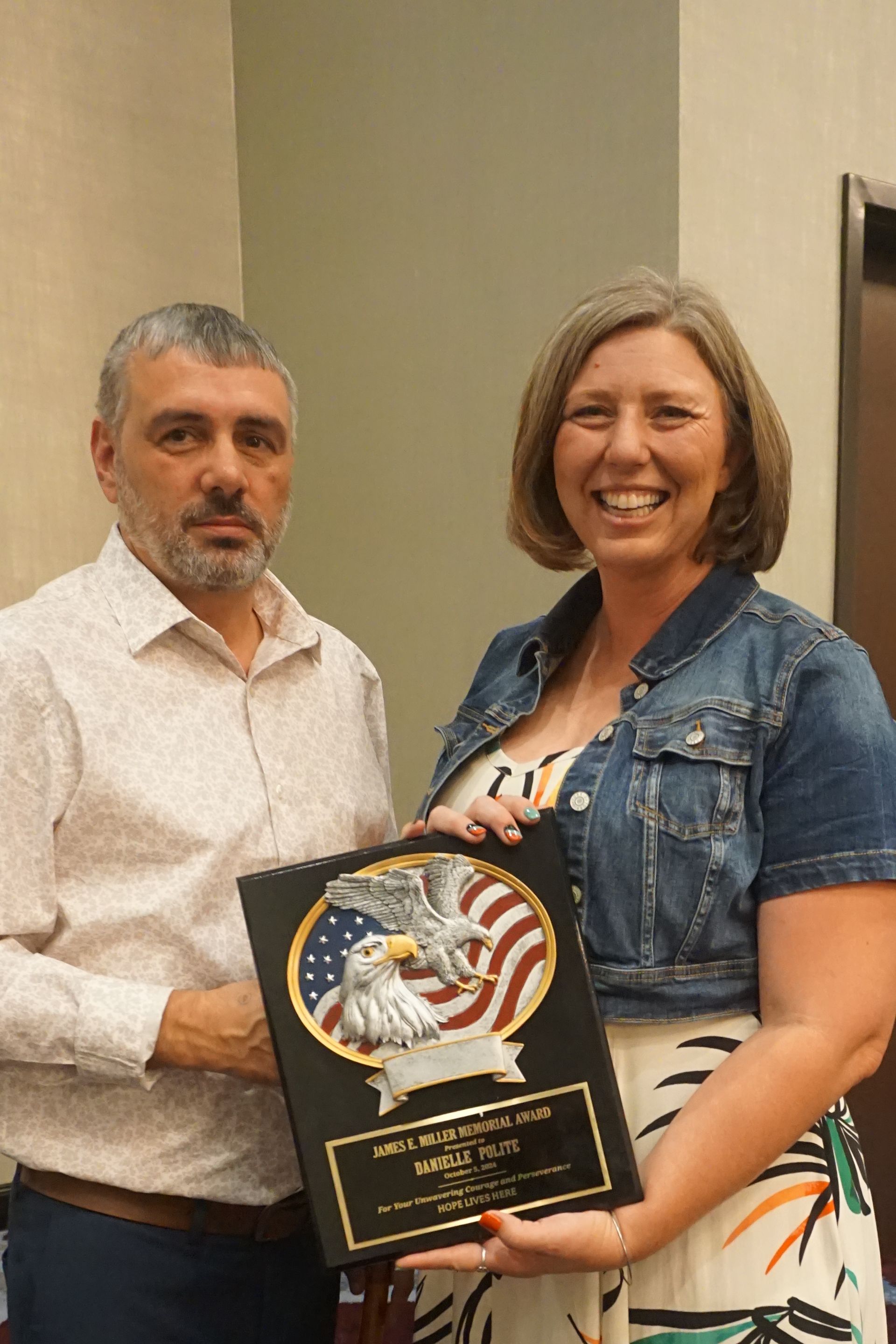 A man and a woman smiling while holding an award plaque featuring an eagle and American flag design.