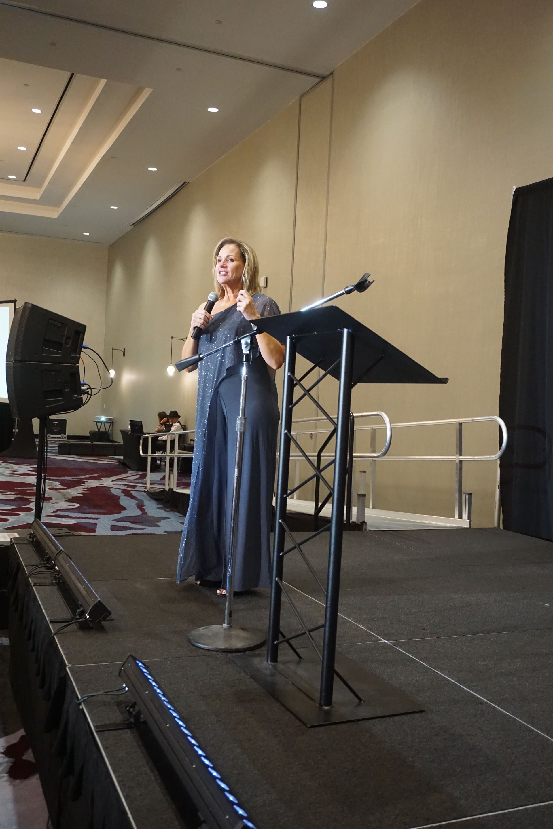 A speaker in a long, dark dress stands at a podium on a stage in a large, modern conference room, holding a microphone.