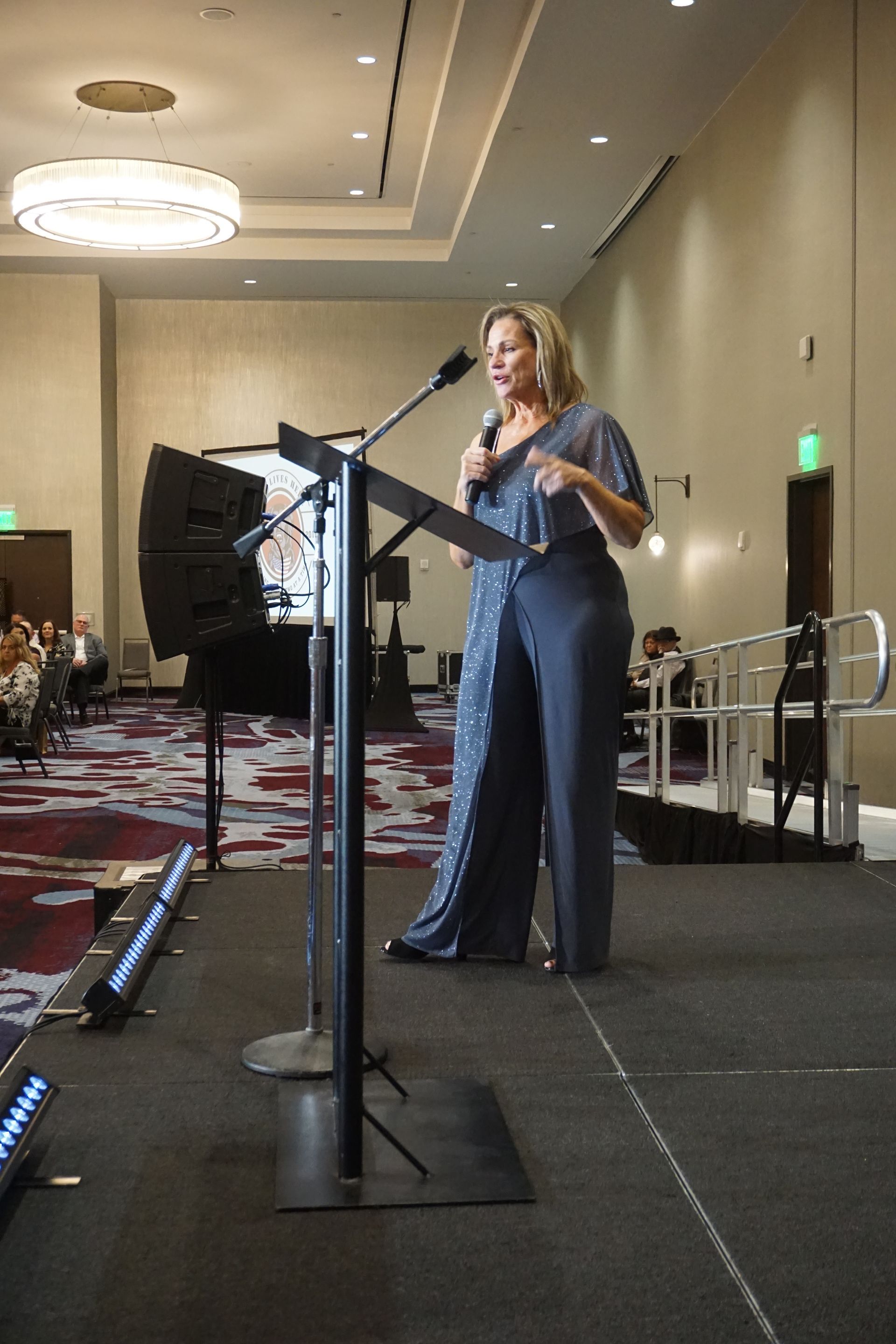 A speaker in a dark jumpsuit stands at a podium on a stage in a conference room, gesturing while addressing an audience.