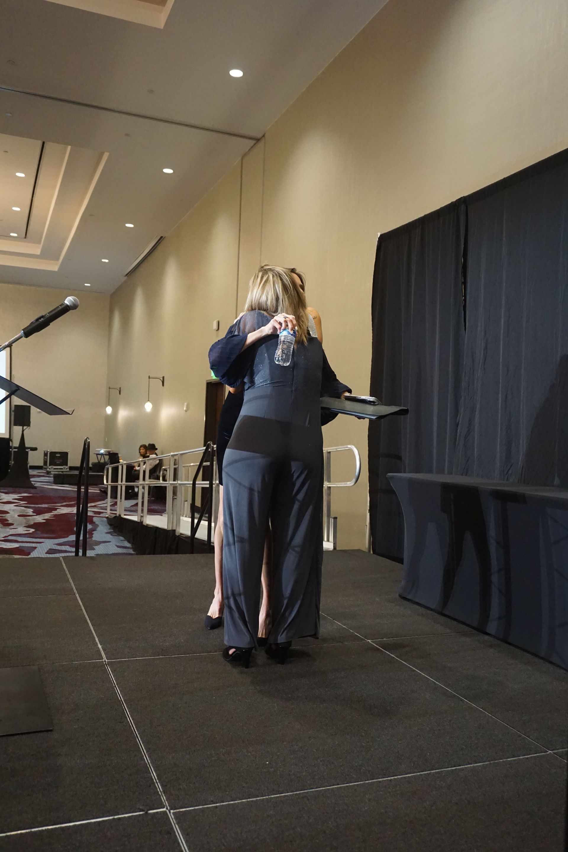 Two people in formal attire embrace on a stage during an event in a conference room.