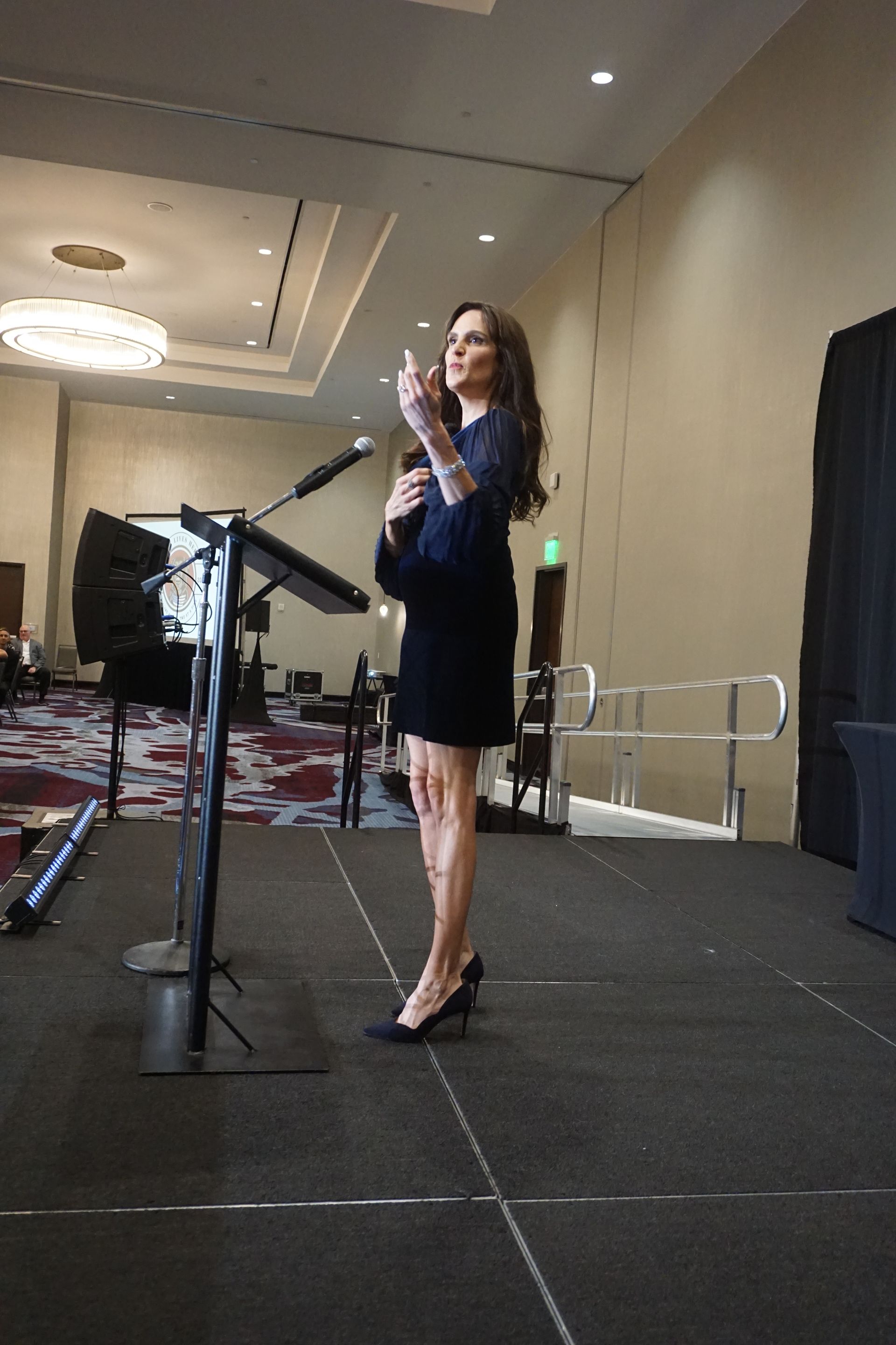 A person in a dark dress speaks at a podium on a stage in a conference room, gesturing with their hand.