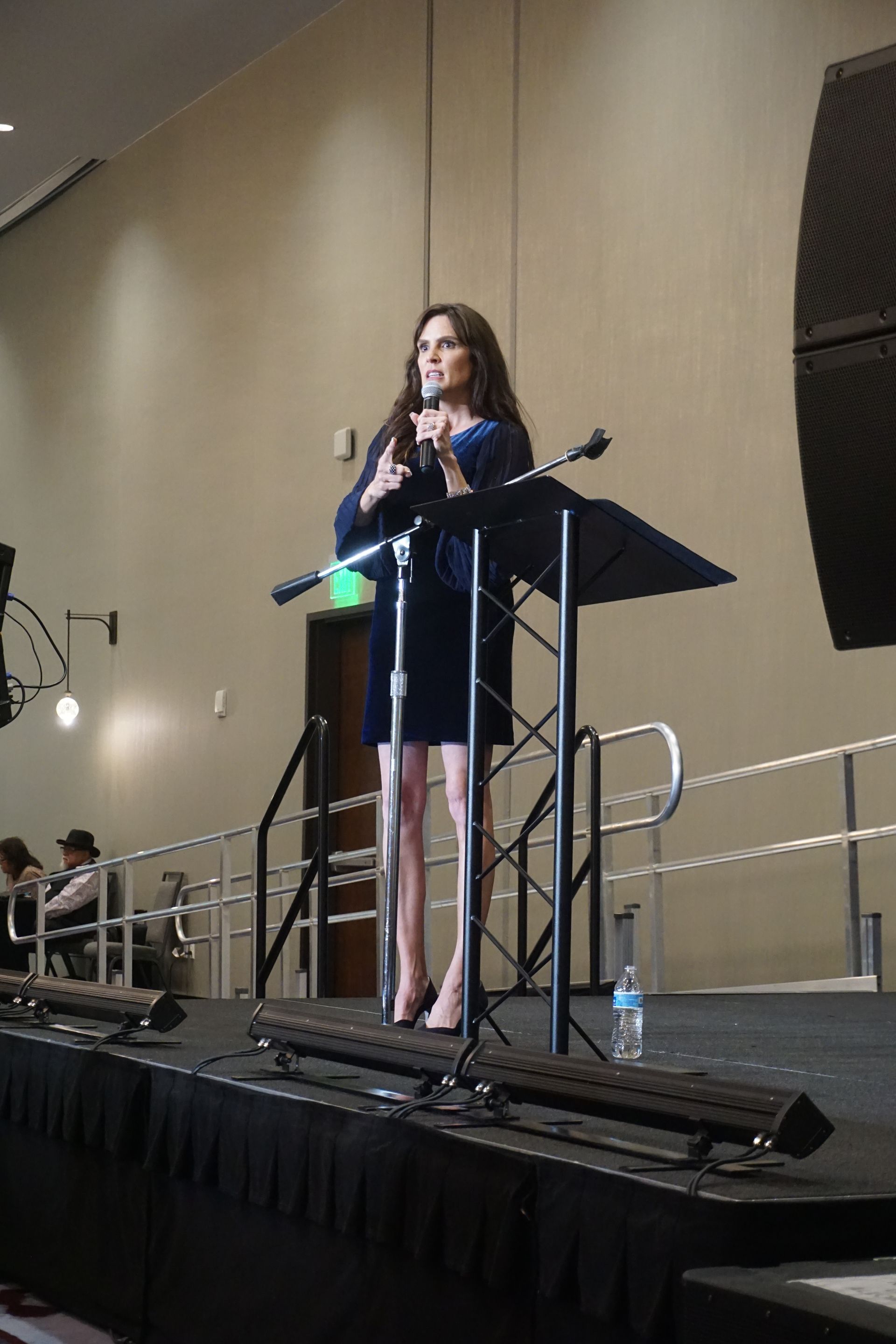 A person stands at a podium on a stage, speaking into a microphone in a brightly lit indoor venue.