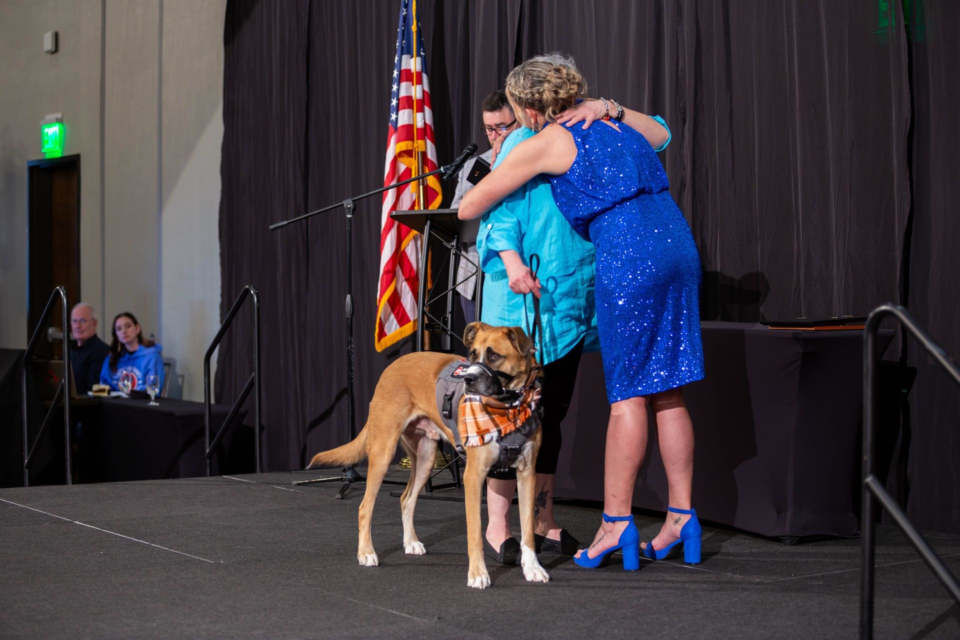 A person in a blue sequined dress hugs another person holding a dog on a stage with an American flag in the background.