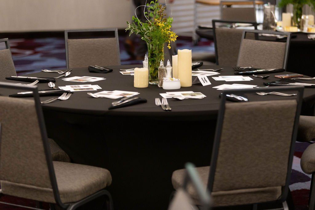 A round event table with a black tablecloth, set with chairs, a central floral arrangement, and pillar candles.