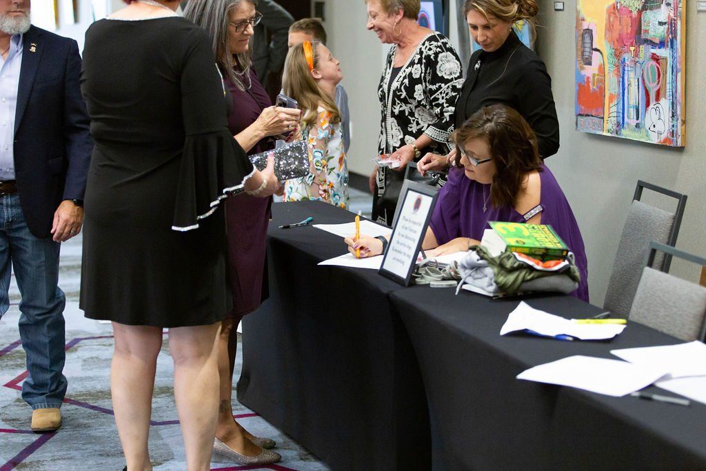 People gather around a table with a black cloth, where a woman signs books in an indoor gallery or event space.