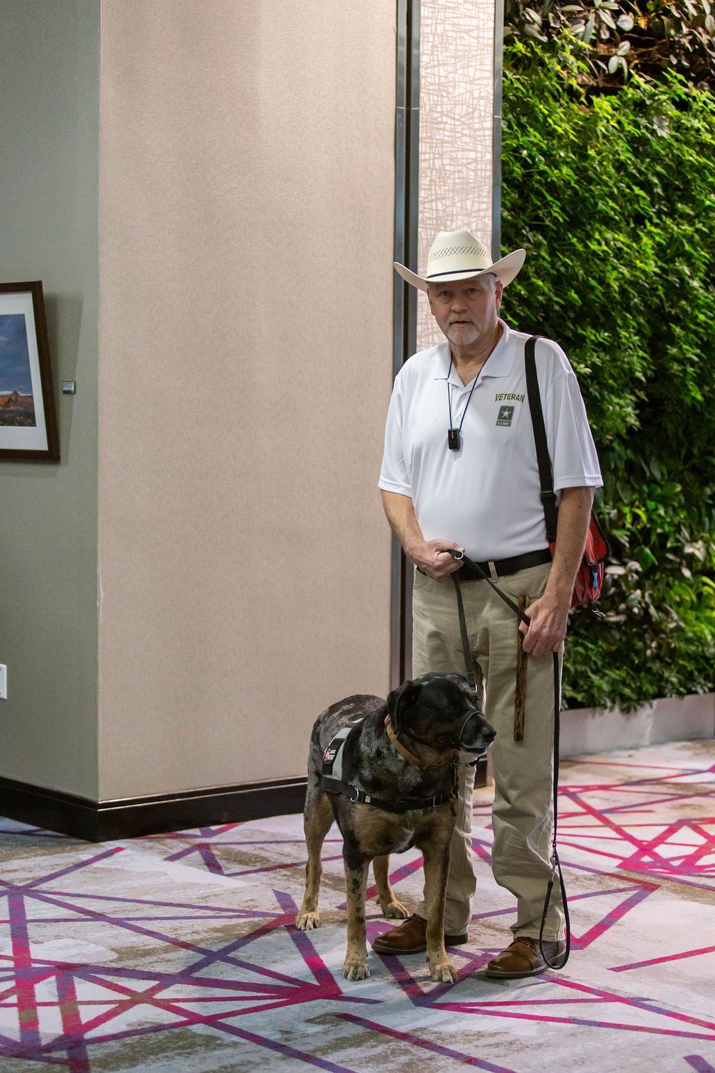 A person in a cowboy hat stands on a patterned carpet next to a service dog in a harness.