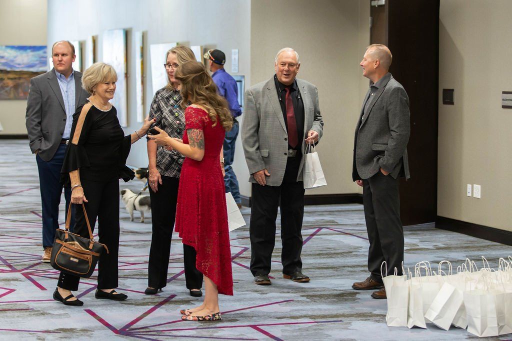 A group of people dressed in formal attire gather and converse in a brightly lit hallway with patterned carpeting.