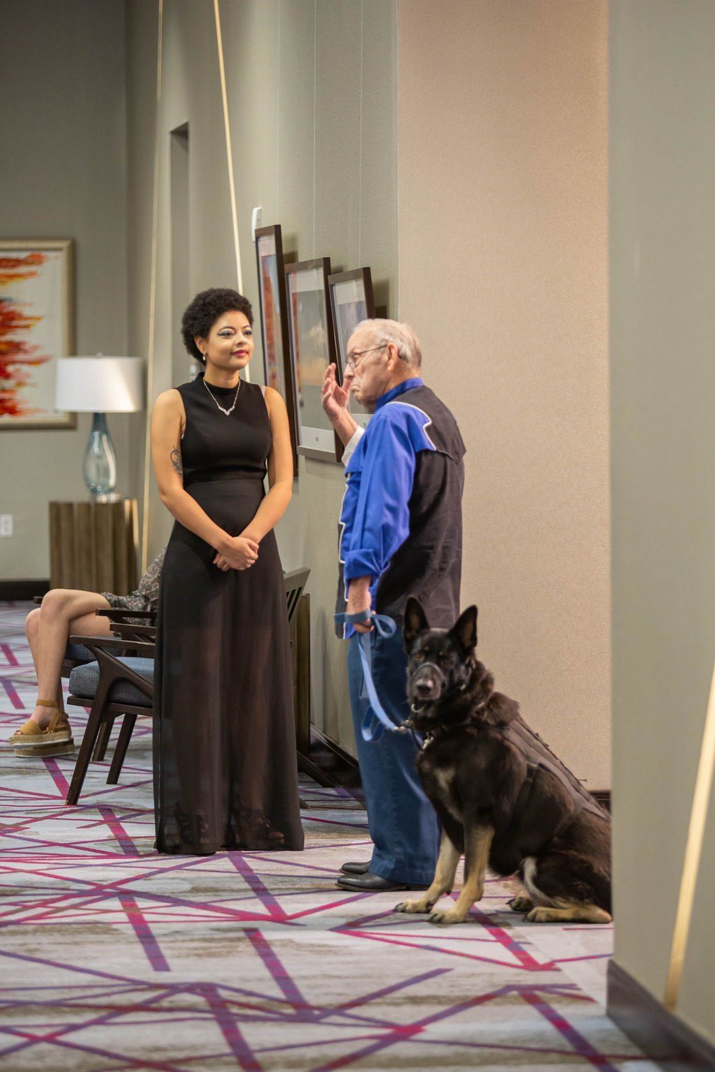 A person in a black dress stands near a person holding a service dog on a leash in a hallway with framed wall art.