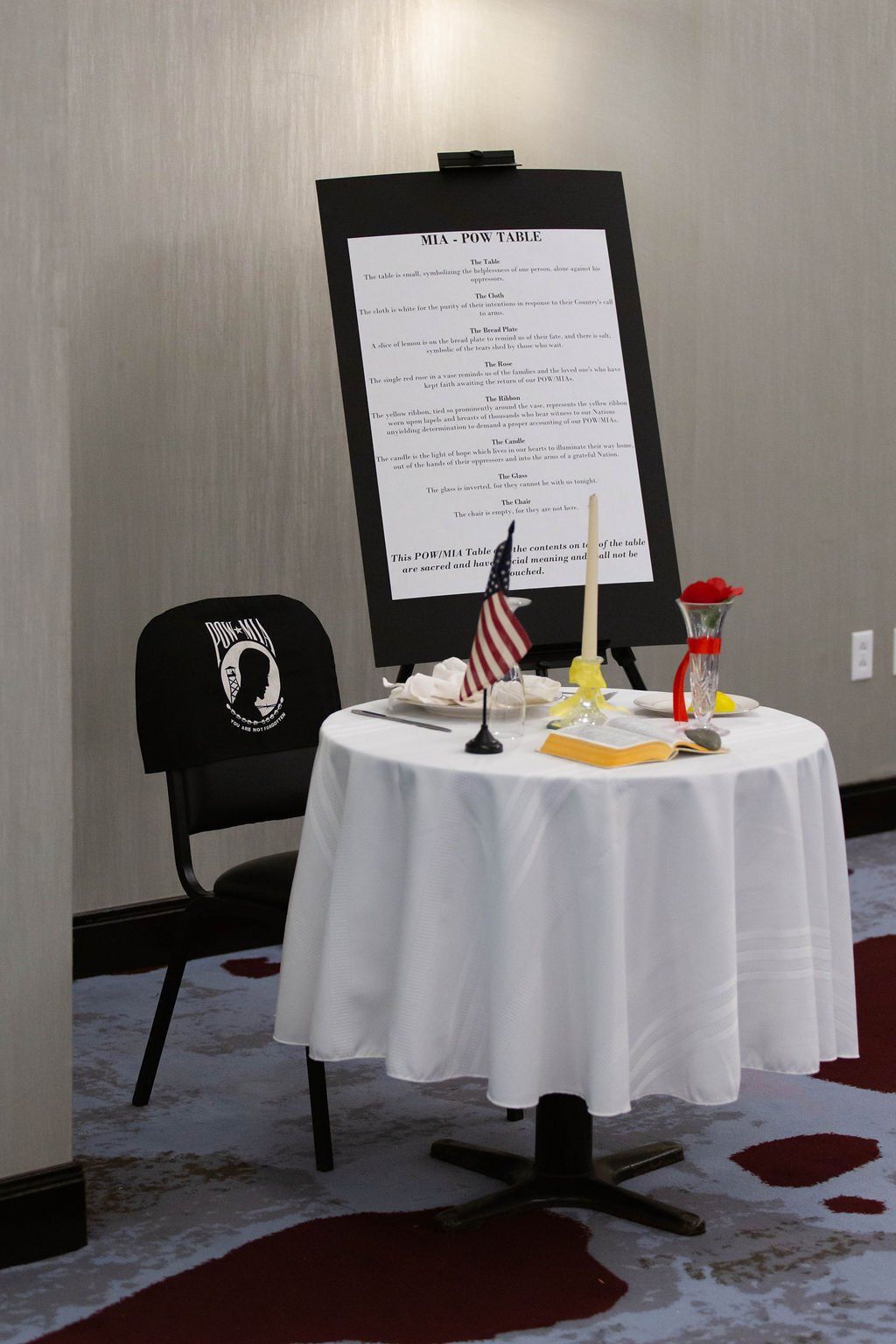 A Missing Man table set with a white cloth, single chair, American flag, candle, rose, and a sign on an easel.
