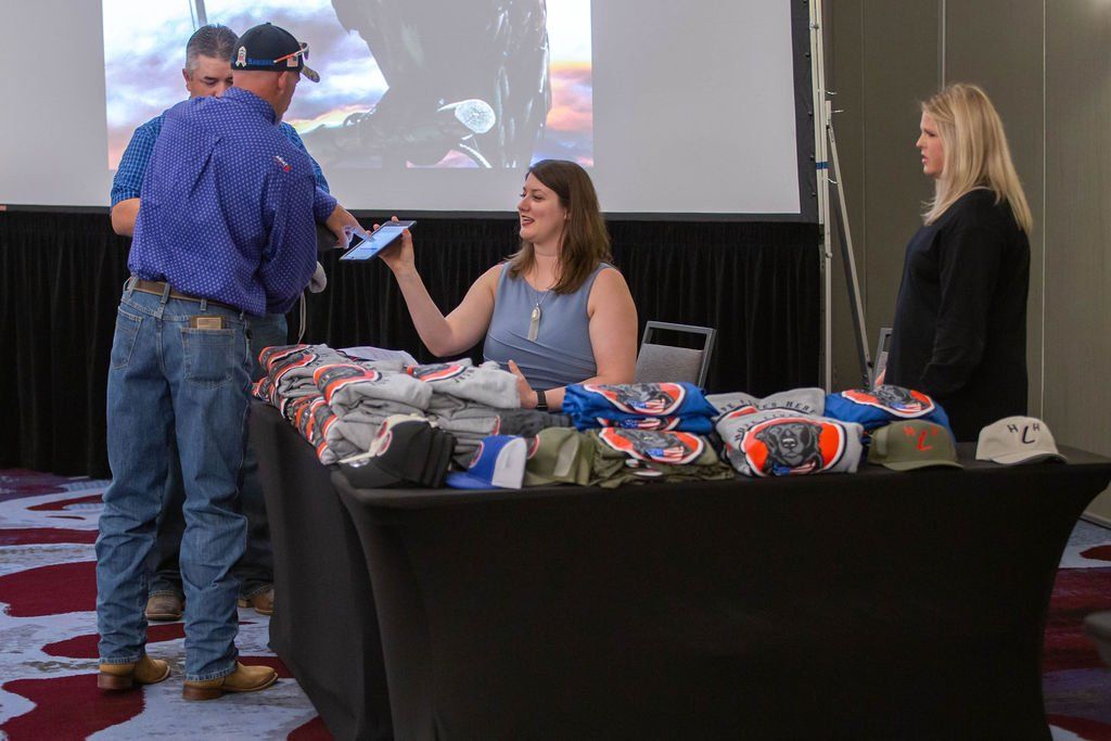 A woman in a blue dress hands a tablet to a man in a blue shirt at a table covered with event merchandise.