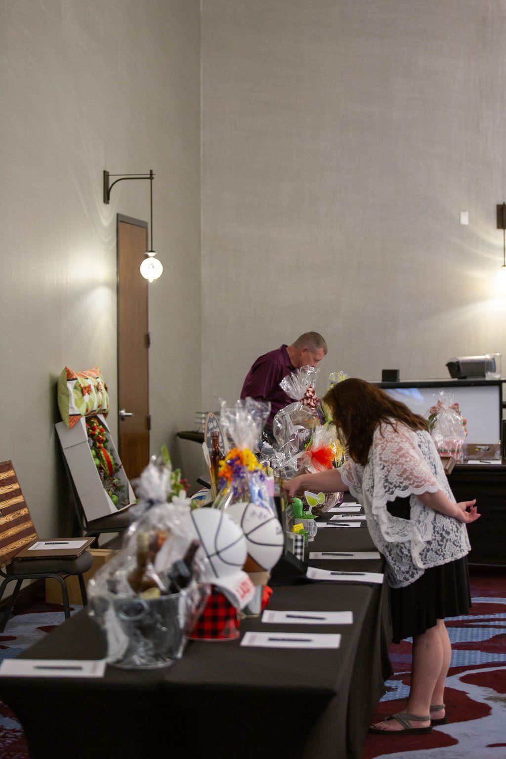 People look at gift baskets arranged on long, black-clothed tables during a silent auction or event in a hall.