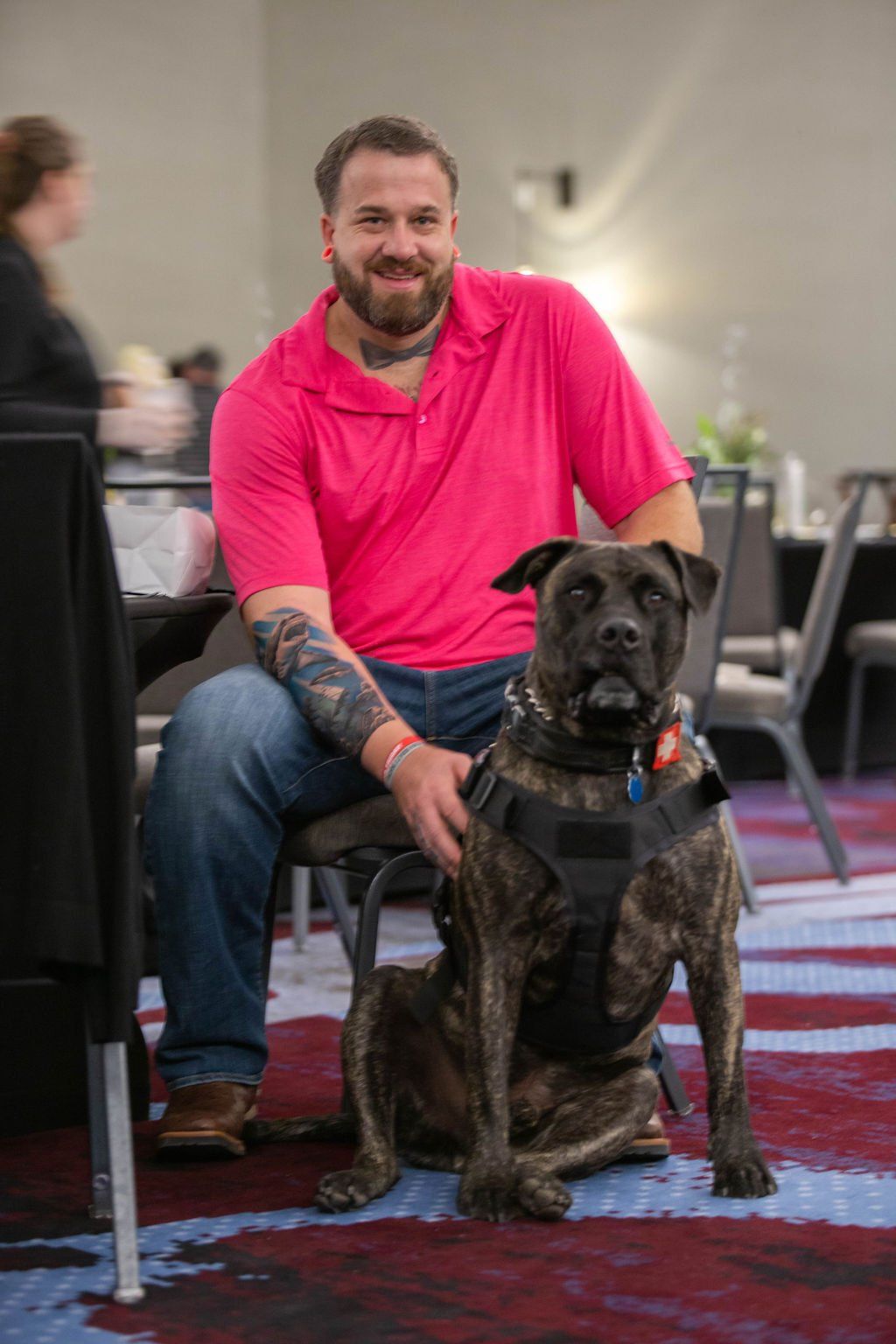 A bearded person in a bright pink polo shirt sits in a chair with their hand resting on a brindle dog wearing a harness.