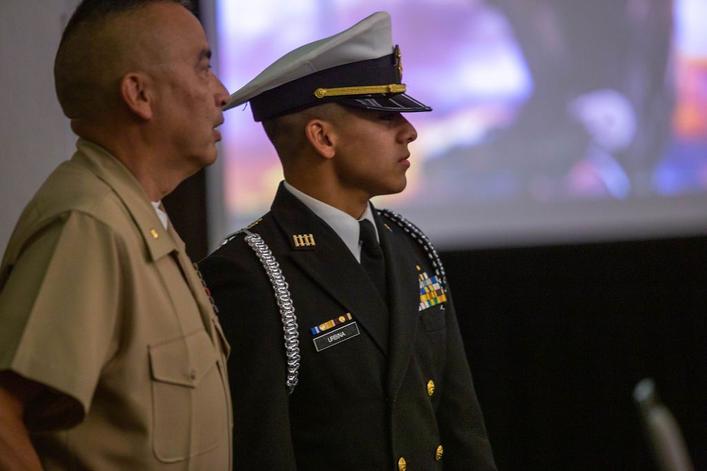 Two military personnel in uniform stand side-by-side, facing forward in an indoor setting.