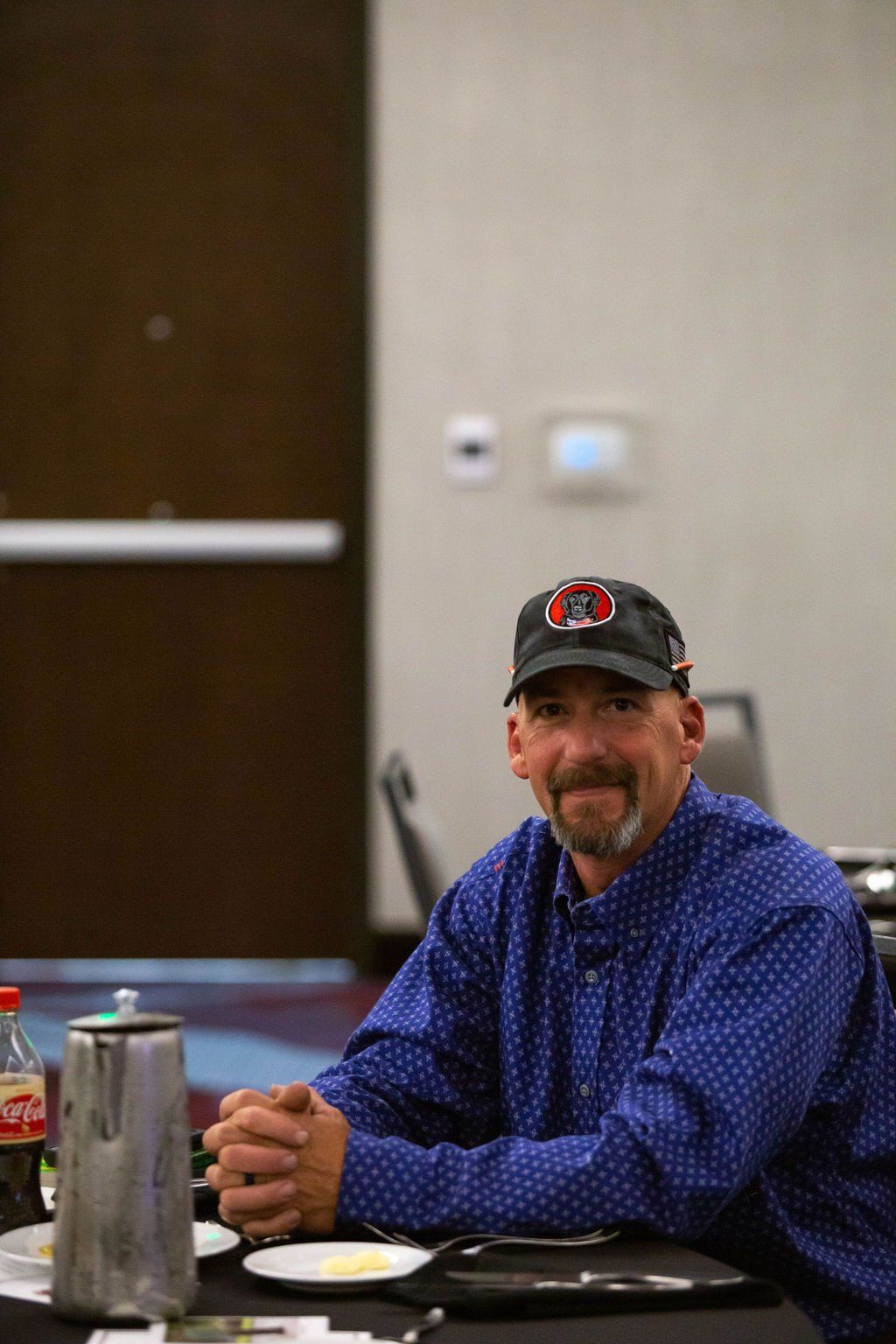 A person wearing a blue patterned shirt and a dark cap sits at a table with their hands clasped in a conference room.