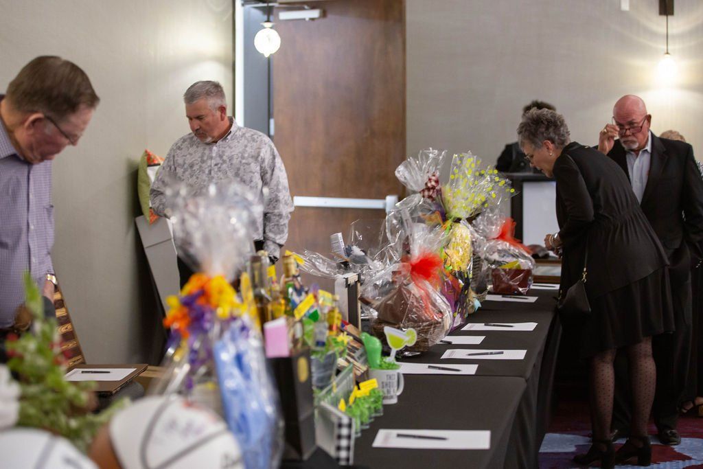 People gather around a long table covered in cellophane-wrapped gift baskets for a silent auction.